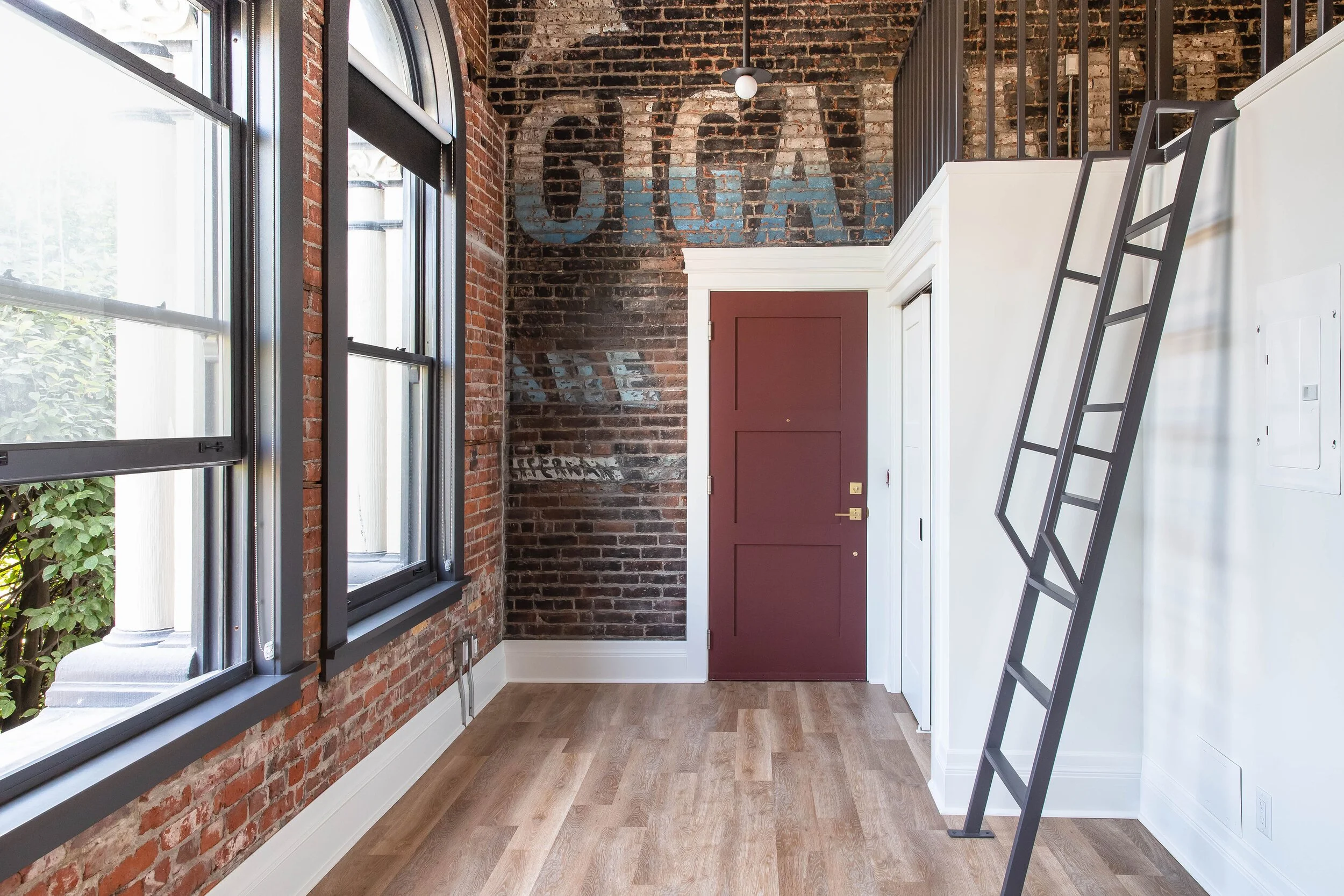 Interior of a room with brick walls, large windows, a red door, a white cabinet, and a black ladder leaning against the wall.