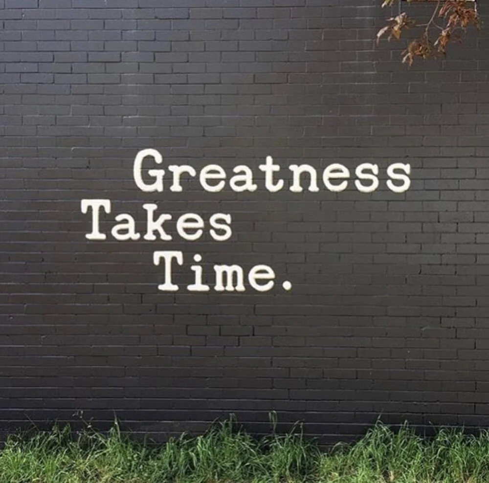 A black brick wall with white painted words: 'Greatness Takes Time' and some grass at the bottom.