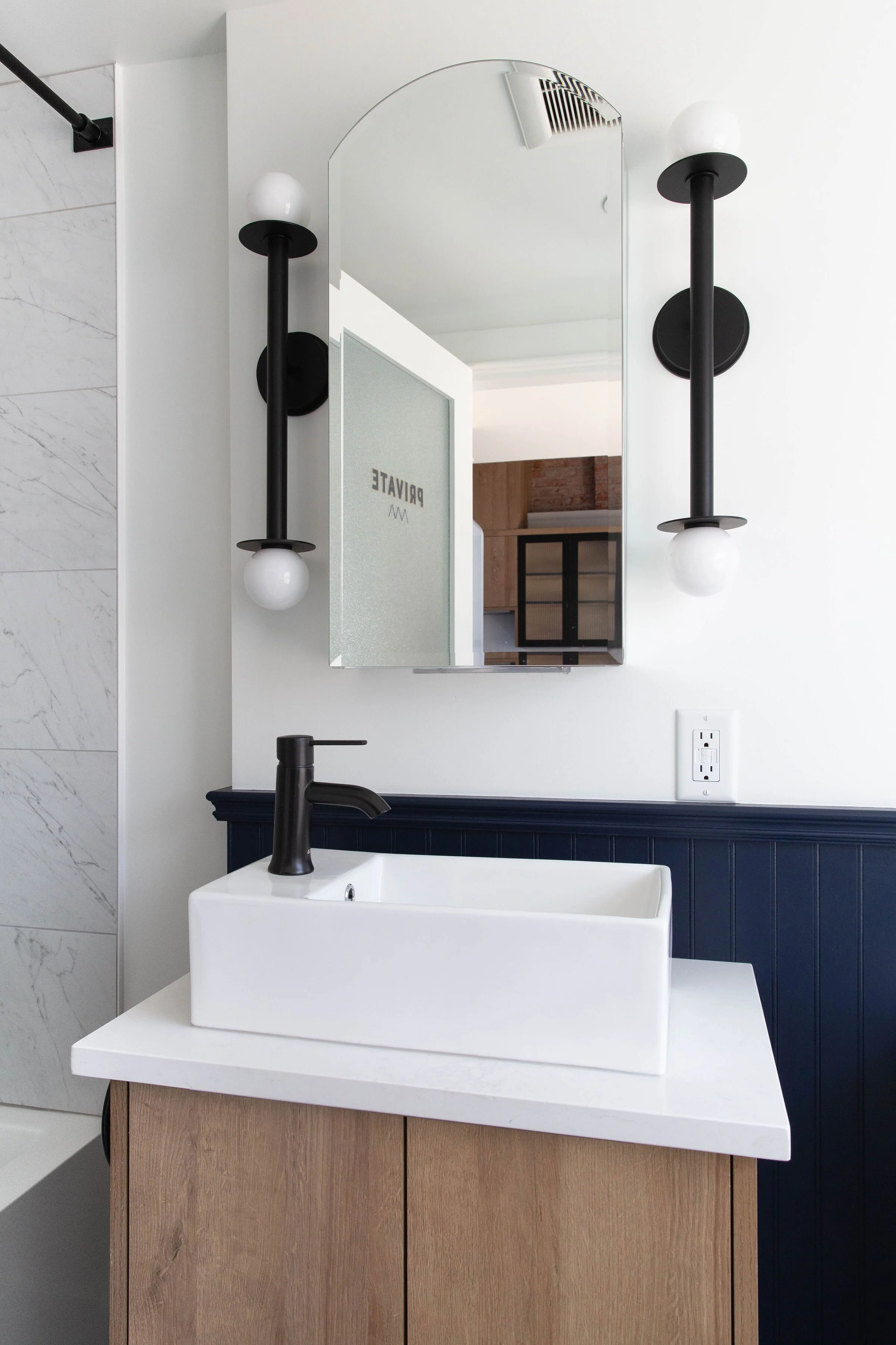 Modern bathroom vanity with a white rectangular vessel sink, black faucet, and a large mirror above. Two black wall-mounted light fixtures with white globes on either side of the mirror. White wall with a dark blue beadboard panel and a power outlet.
