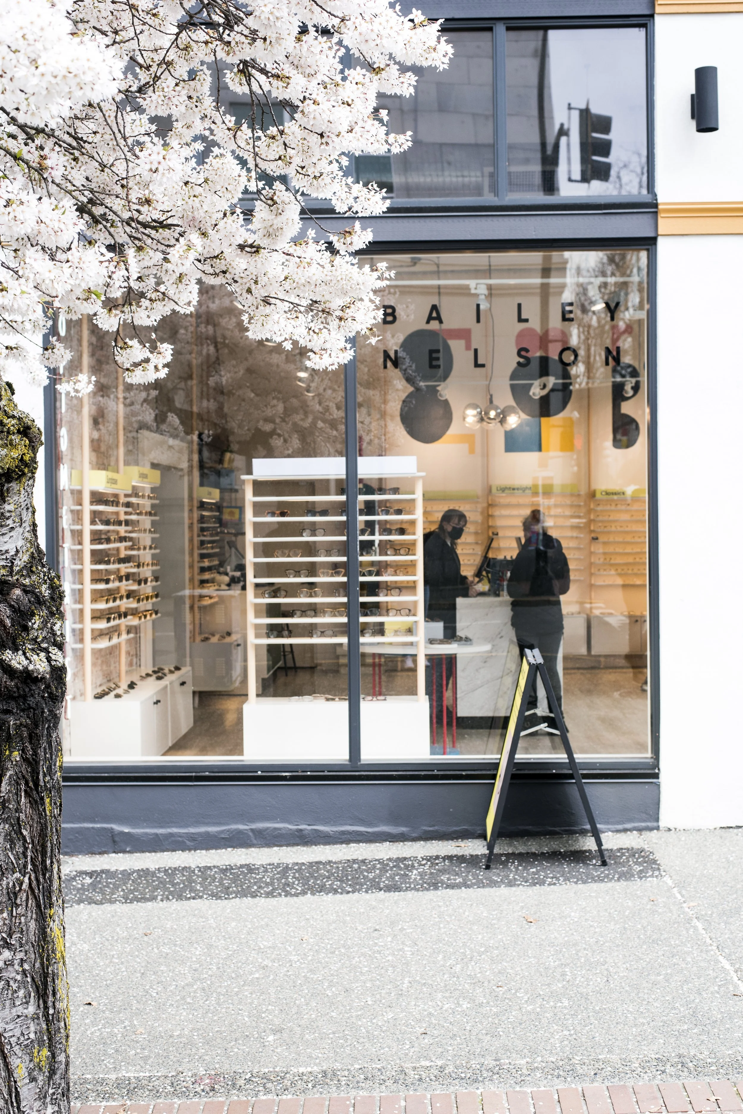Storefront with large glass window displaying eyeglasses, sign reading 'BAILEY NELSON', pink flowering tree in foreground, sidewalk outside, two people inside.