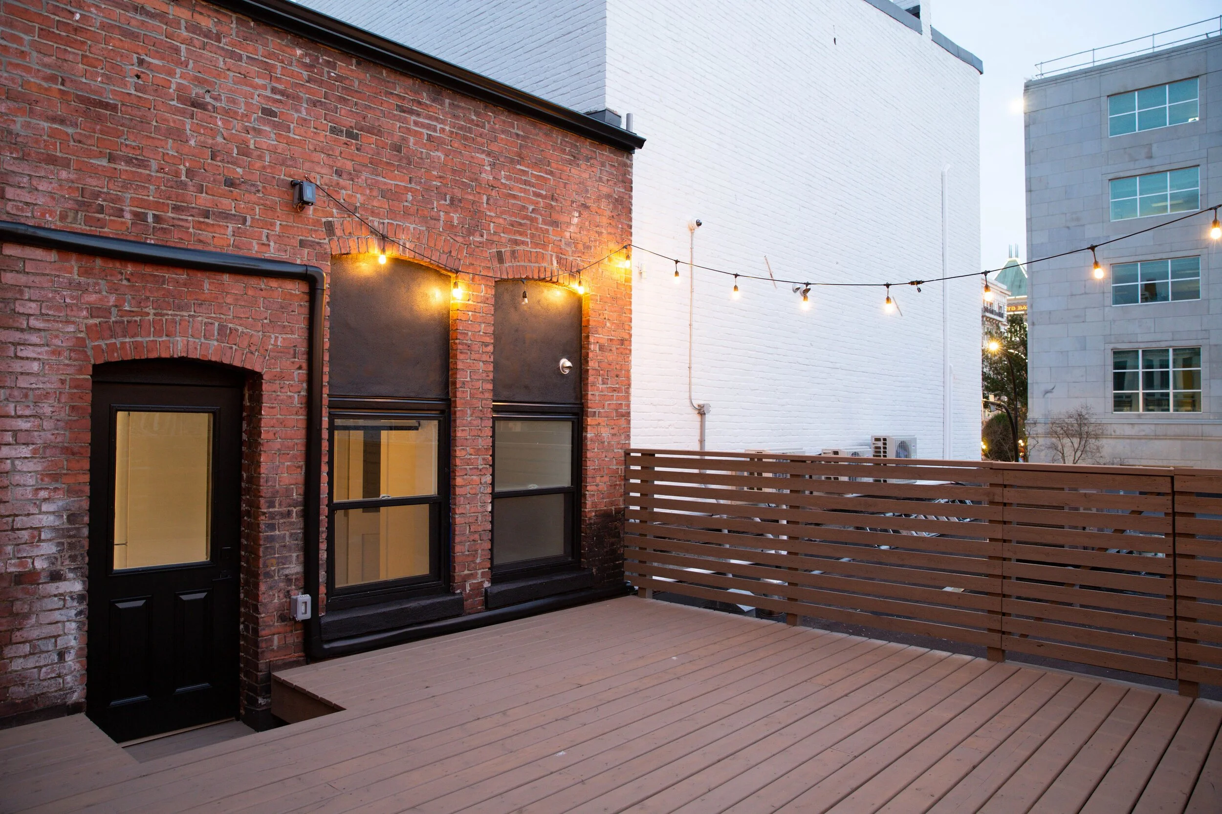 Urban rooftop patio with wooden deck, string lights, brick and white wall buildings in the background.