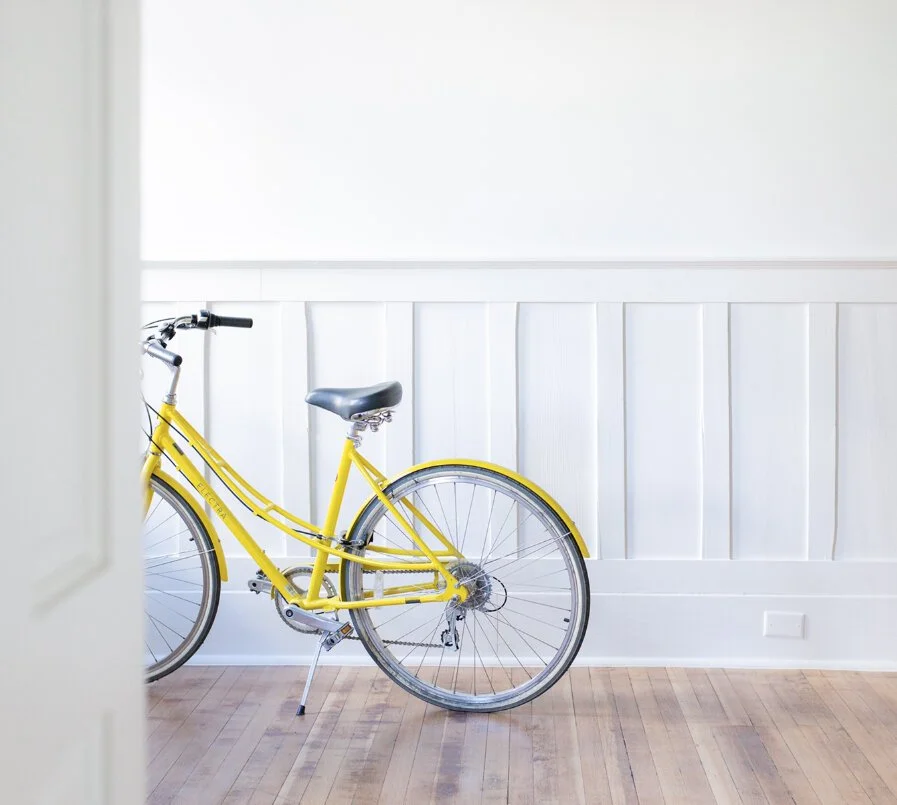 A yellow bicycle with a black seat parked indoors against a white wall with wood paneling and hardwood floor.