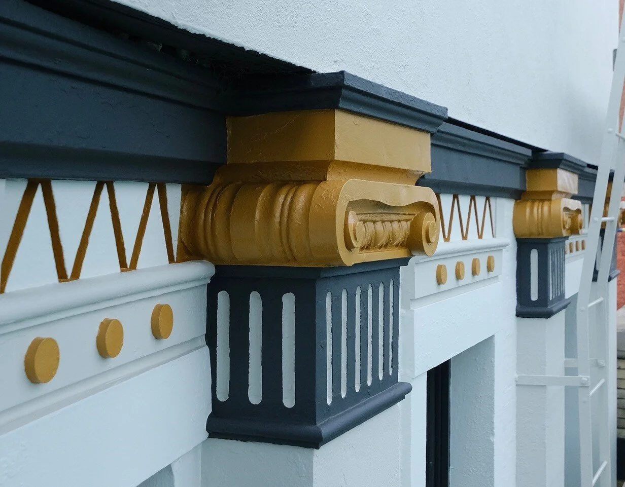 Close-up of decorative architectural molding on a building's exterior, featuring gold and black painted wooden designs with white background.