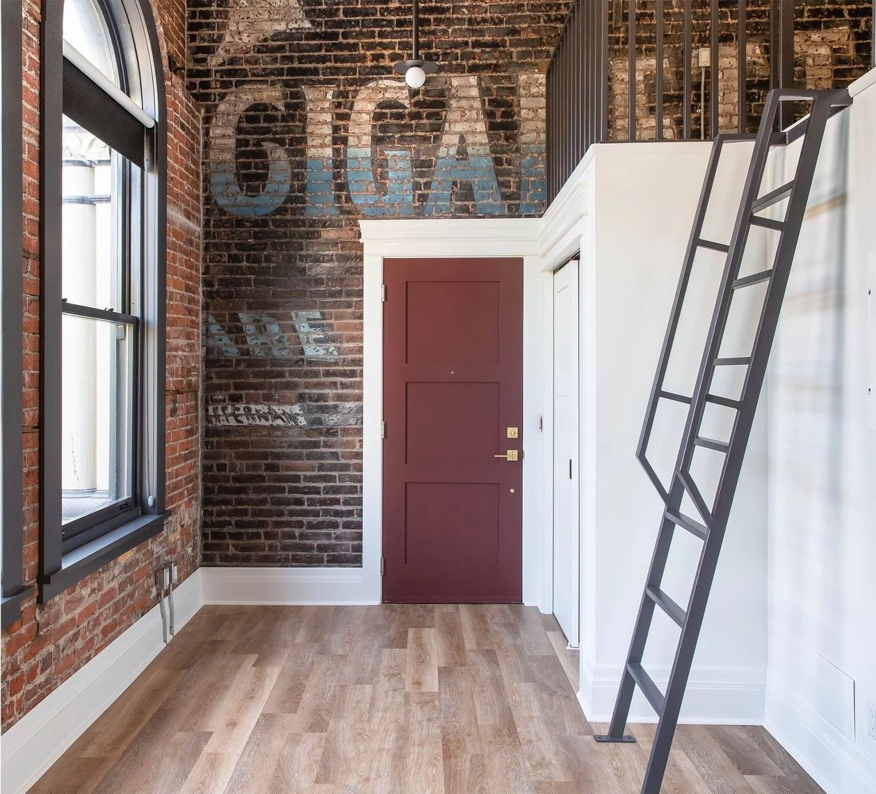 Interior of a loft-style apartment with an exposed brick wall, a maroon door, a black ladder leading to a loft, and graffiti on the bricks near the ceiling that spells out 'COVA'.