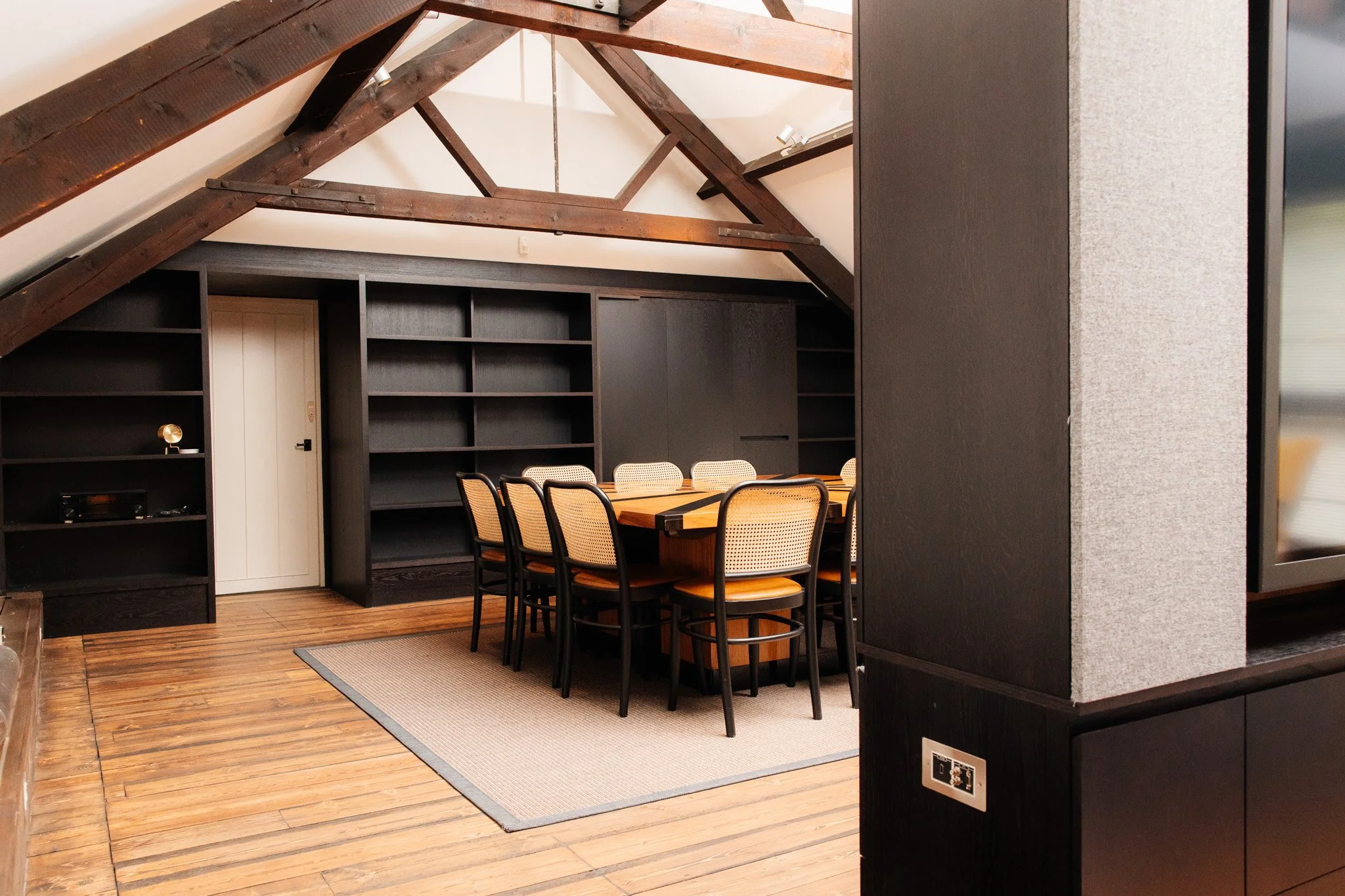 A dining room with a wooden table and six chairs under a sloped ceiling with dark wooden beams, black shelves on the wall, and a beige rug on wooden flooring.