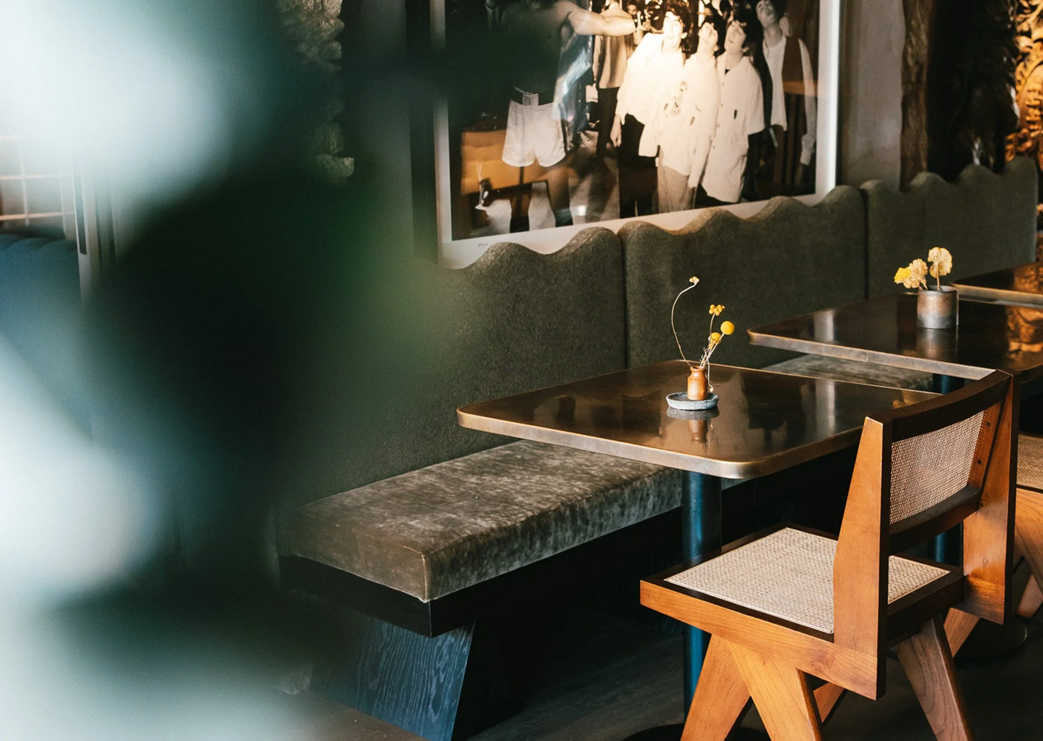 Empty restaurant table with a small vase of yellow flowers, a wooden chair, and a black dining bench, with artwork of a black-and-white photograph on the wall in the background.