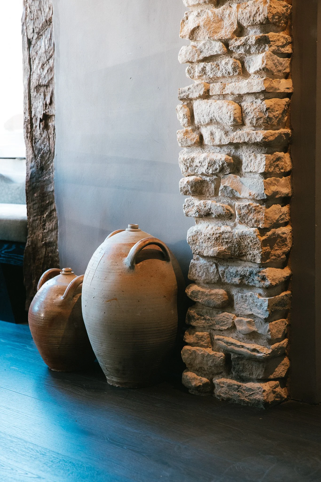 Two large brown ceramic vases with handles, placed on black wooden floor next to a rustic stone column and a gray wall.