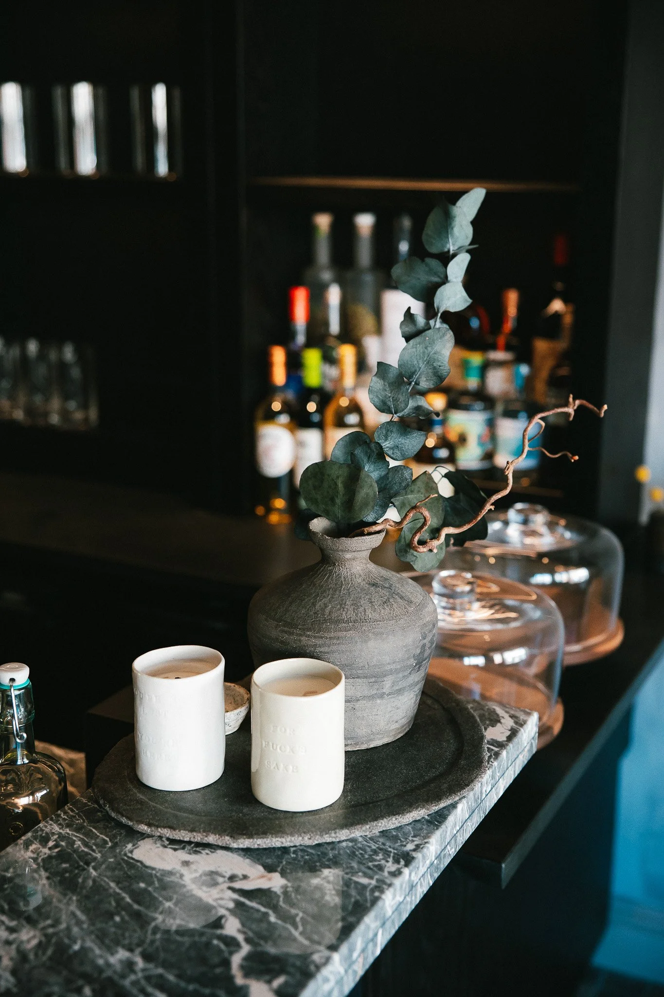 A marble counter with a gray vase holding eucalyptus branches and two white candles, with a blurred bar and liquor bottles in the background.