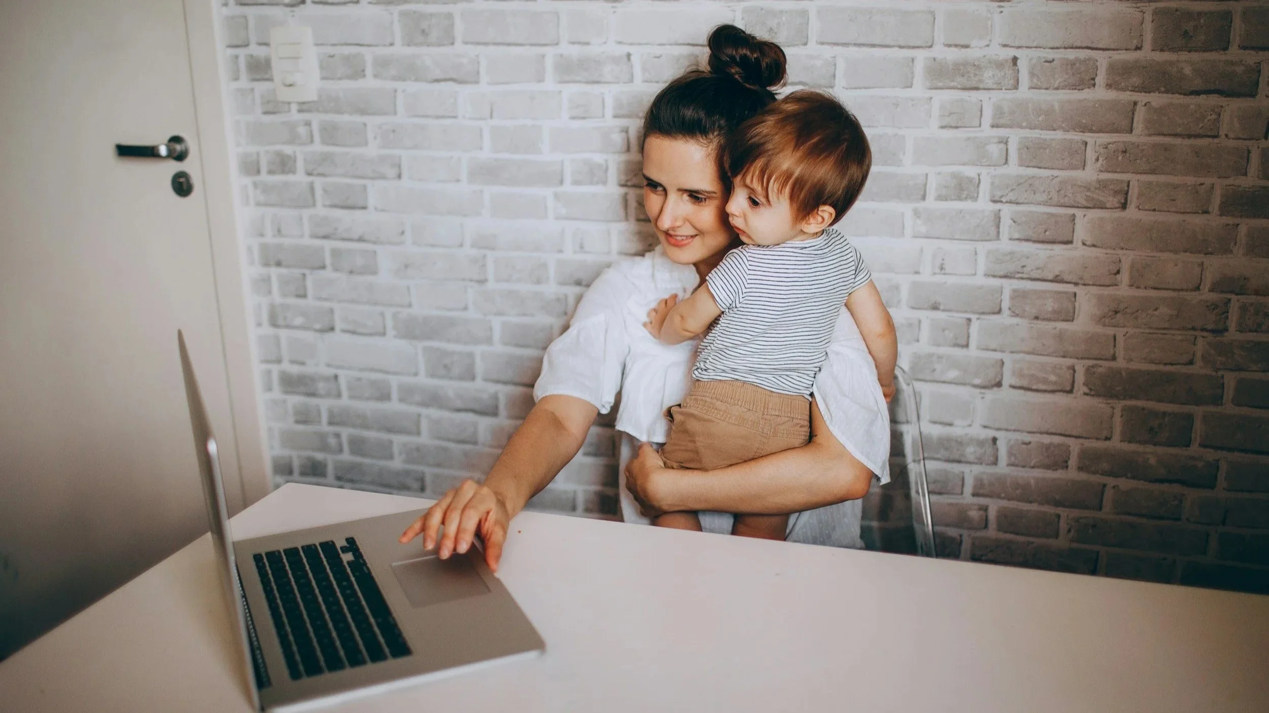 Una mujer joven con su cabello en un chongo, sosteniendo a un niño pequeño, ambos viendo y sonriendo frente a una computadora portátil en una mesa, con un fondo de pared de ladrillos de color gris claro.