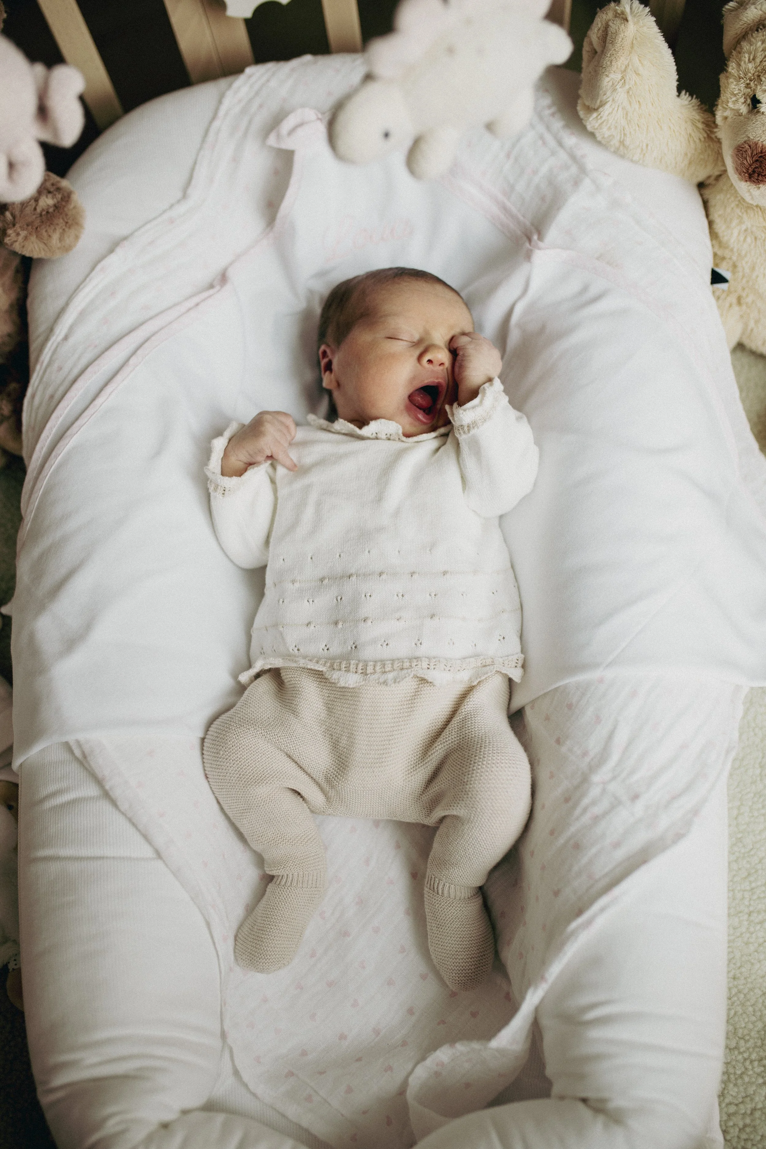 A baby sleeping in a white bassinet surrounded by plush teddy bears, yawning with one hand near their face.