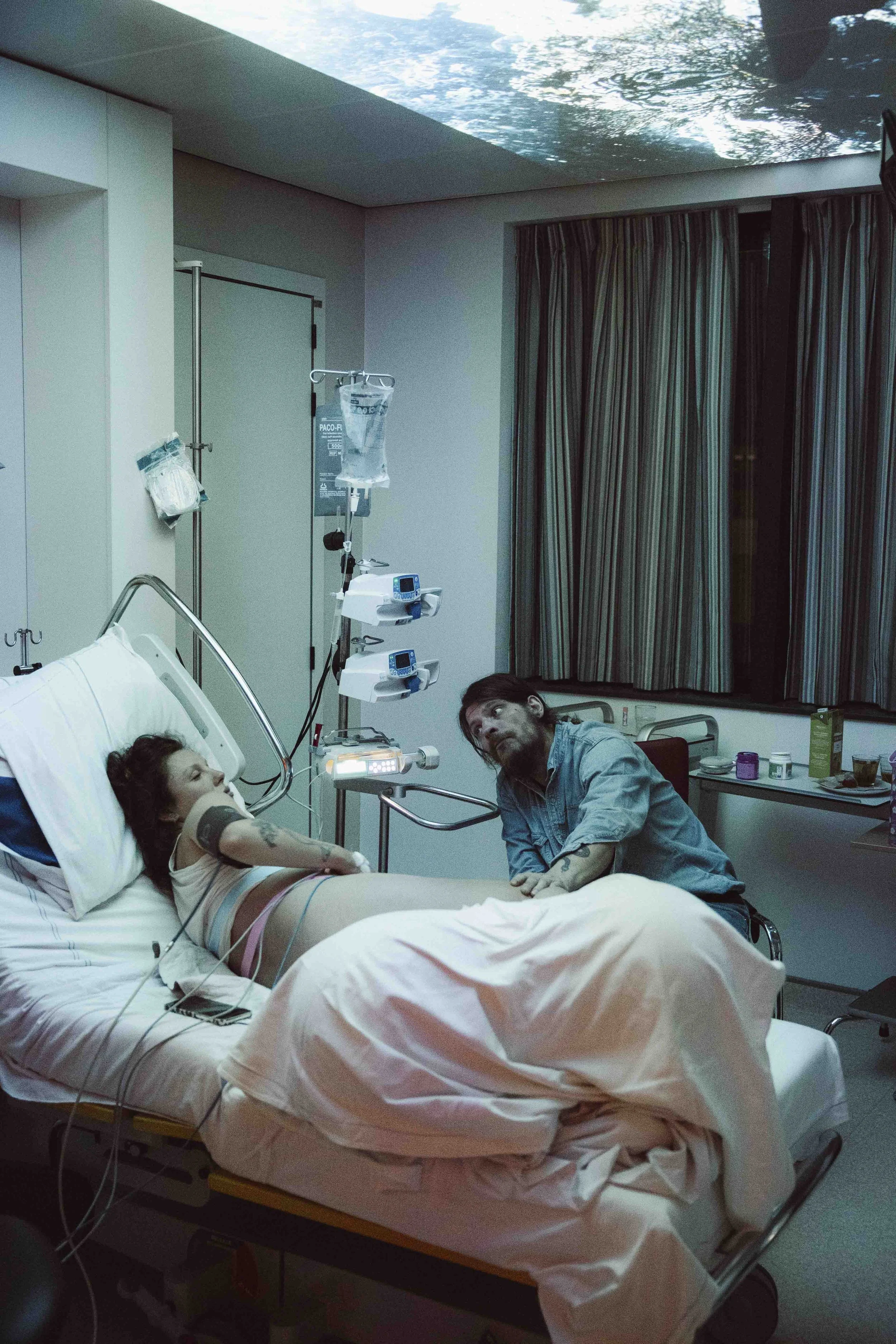 Een vrouw ligt in een ziekenhuisbed met medische lijnen en apparaten, terwijl een man naast haar zit en met haar praat. Er zijn medische apparatuur en een raam met gordijnen op de achtergrond.  Geboortefotografie door Maud Fernhout (de Kraamfotograaf