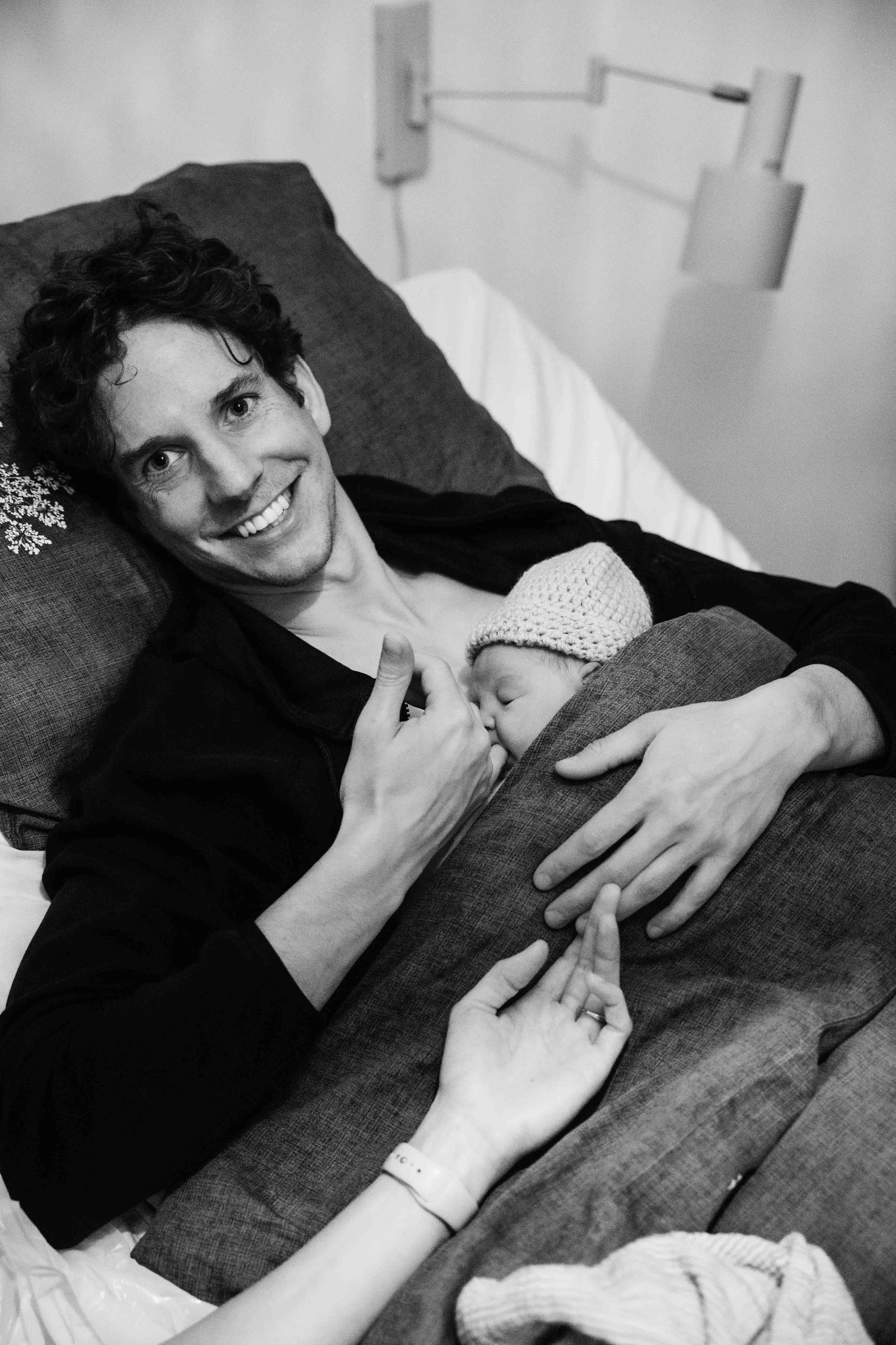 Een jonge vader ligt op een bed na de bevalling met zijn pasgeboren dochter, lacht en wijst met zijn duim omhoog.  Geboortefotografie door Maud Fernhout (de Kraamfotograaf)