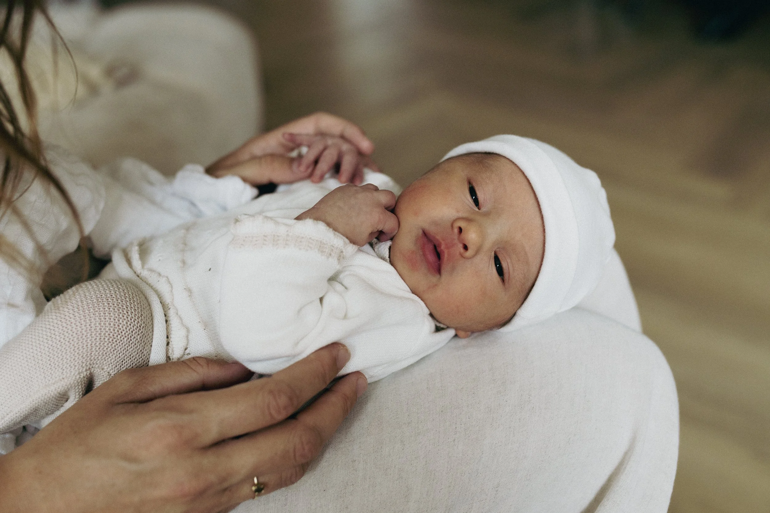 A baby lying on a person's lap, wearing a white outfit and a white hat, with a hand gently touching its chest, in a cozy indoor setting.