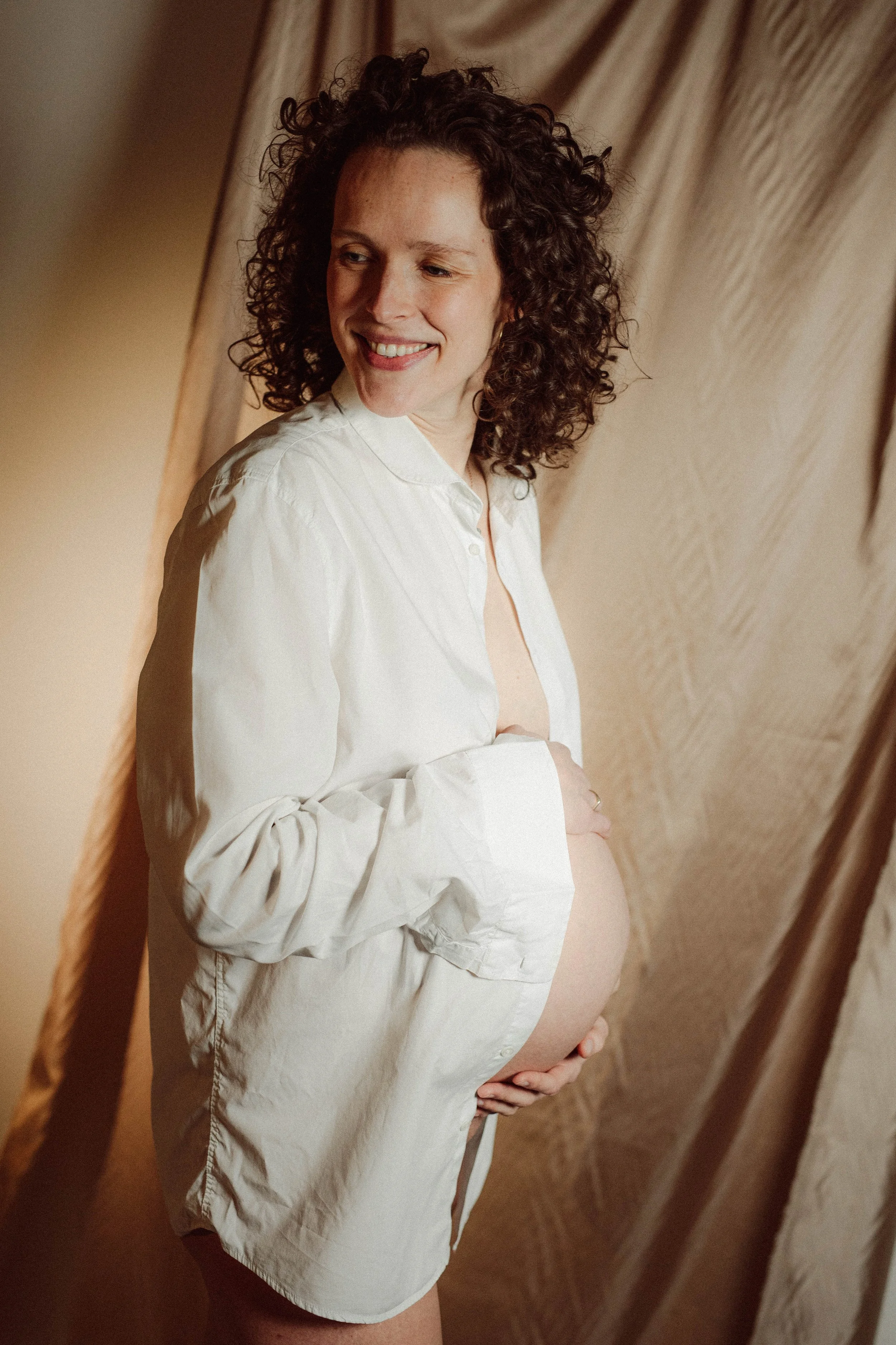 Vrouw met donker, krullend haar en een witte blouse, zittend op een stoel, terwijl ze haar buik vasthoudt.