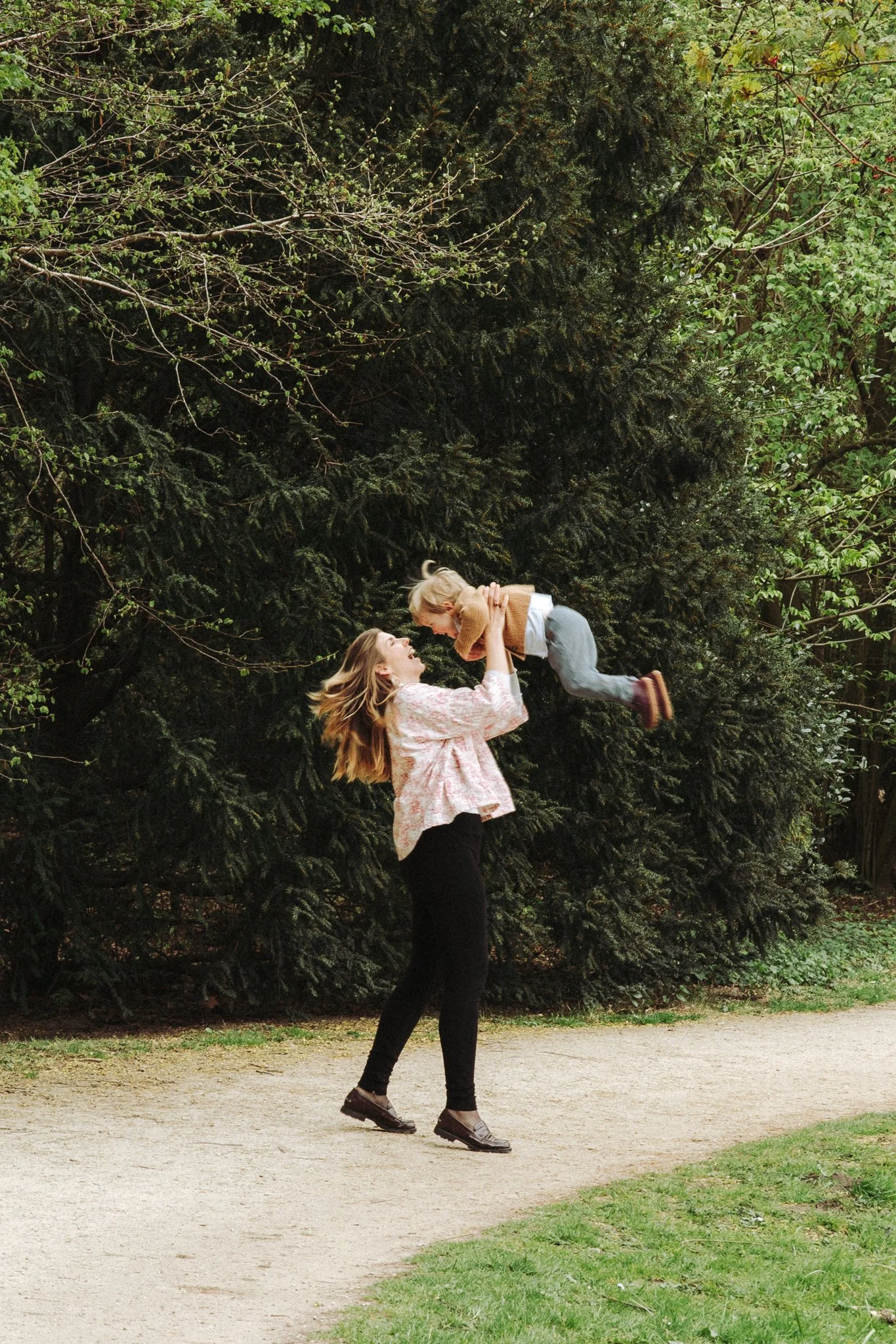 Twee jonge vrouwen die plezier hebben in een park, met een grote groene boom op de achtergrond. Eén vrouw tilt de andere op, die lachend omhoog kijkt.