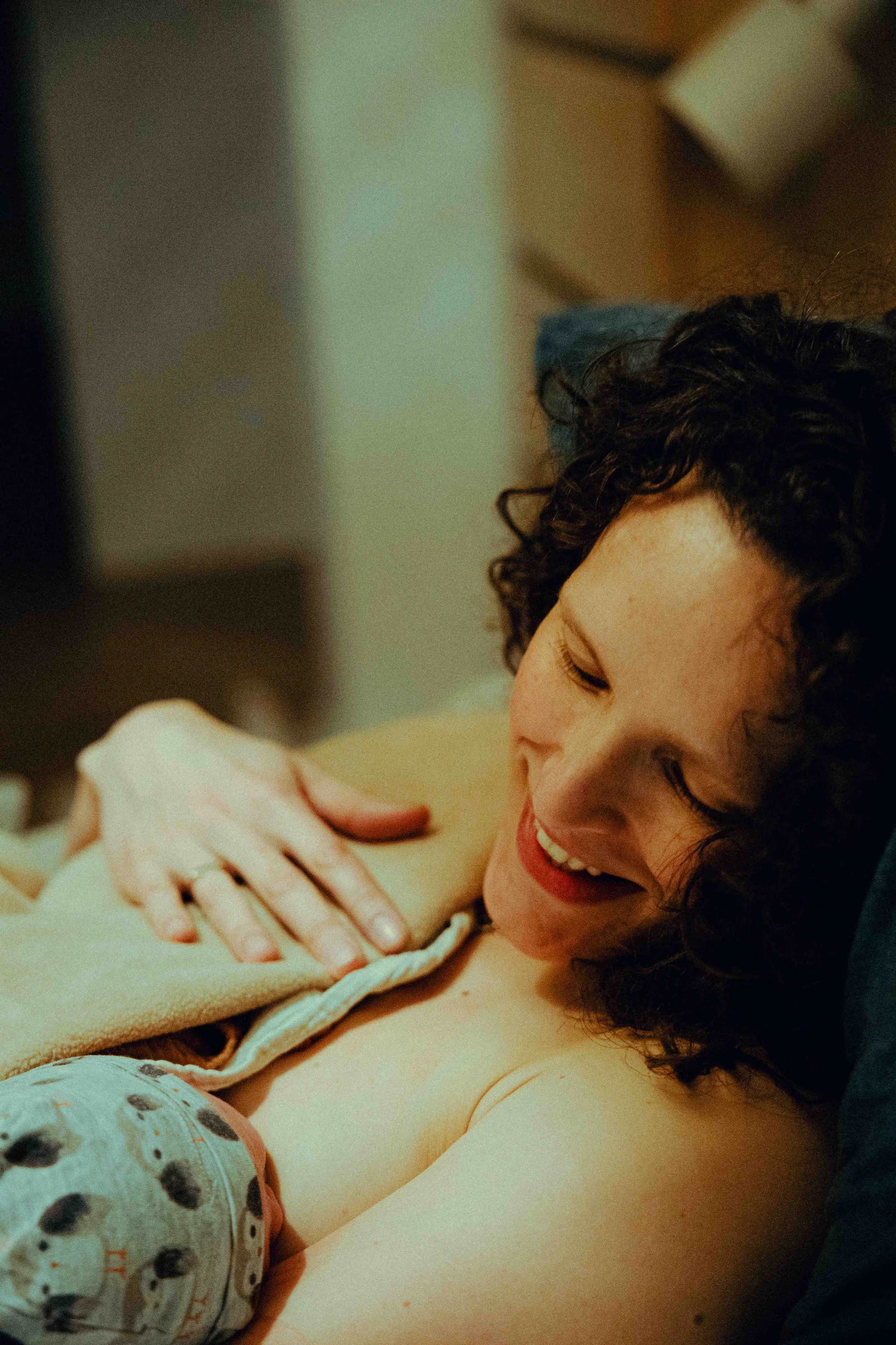 Een jong meisje met krullend haar koestert een baby en glimlacht terwijl ze haar hand op de borst van de baby legt.  Geboortefotografie door Maud Fernhout (de Kraamfotograaf).