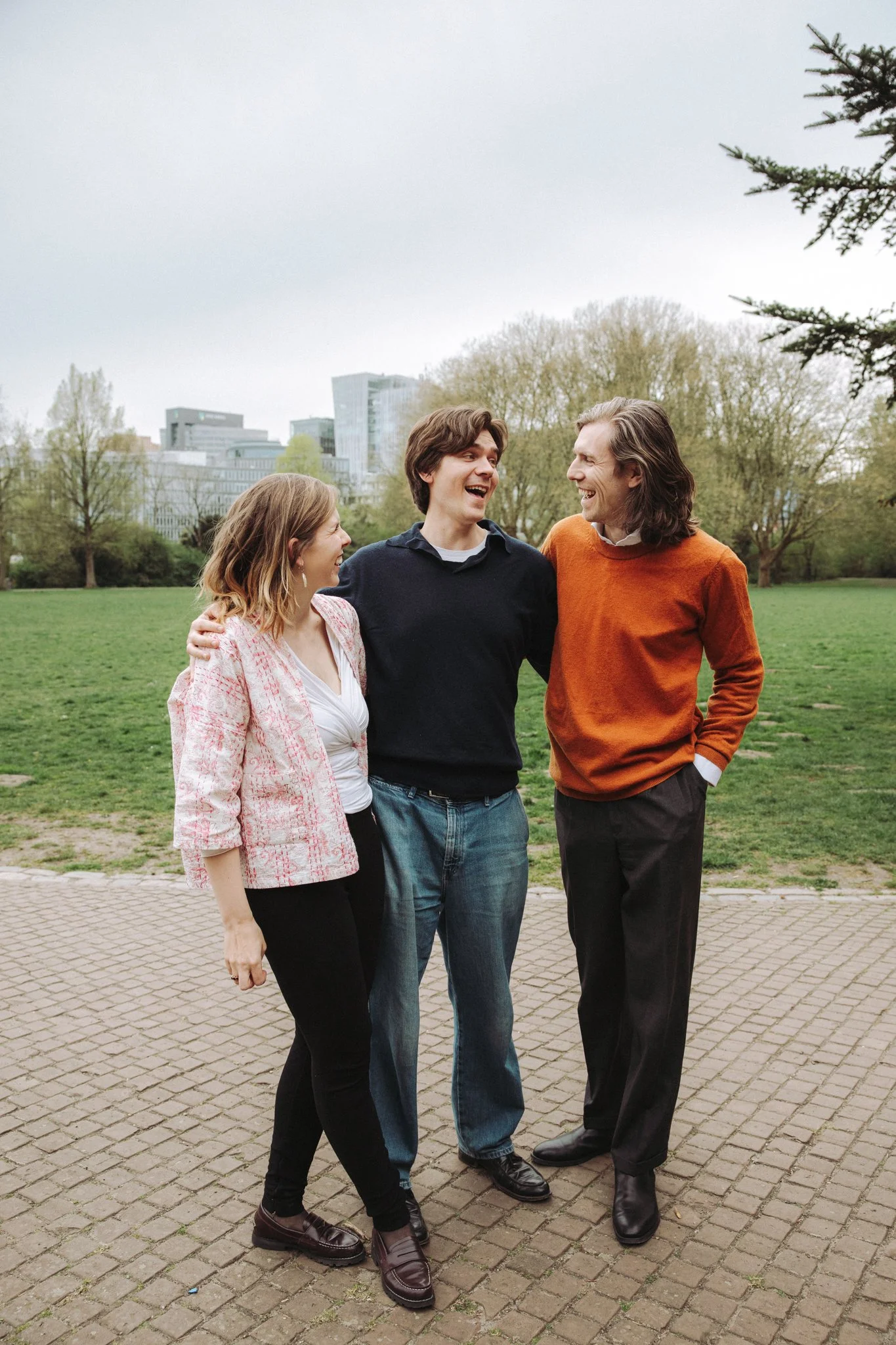 Drie mensen staan in een park, lachen en praten met elkaar, op de achtergrond bomen en moderne gebouwen.