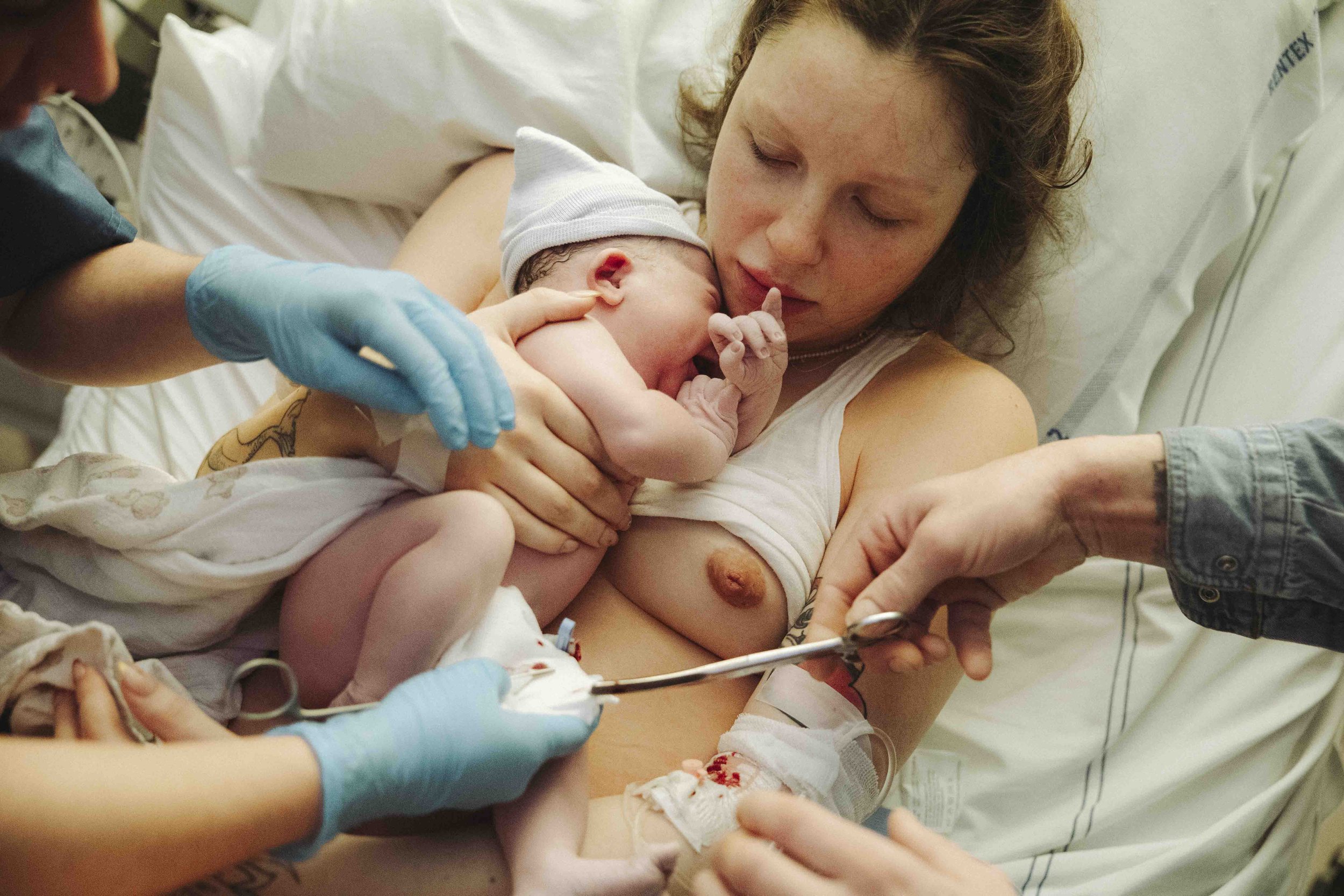 Pasgeboren baby met een mutsje wordt geboren, omringd door medische medewerkers en een vrouw die op de bevaltafel ligt in een ziekenhuis.  Geboortefotografie door Maud Fernhout (de Kraamfotograaf).