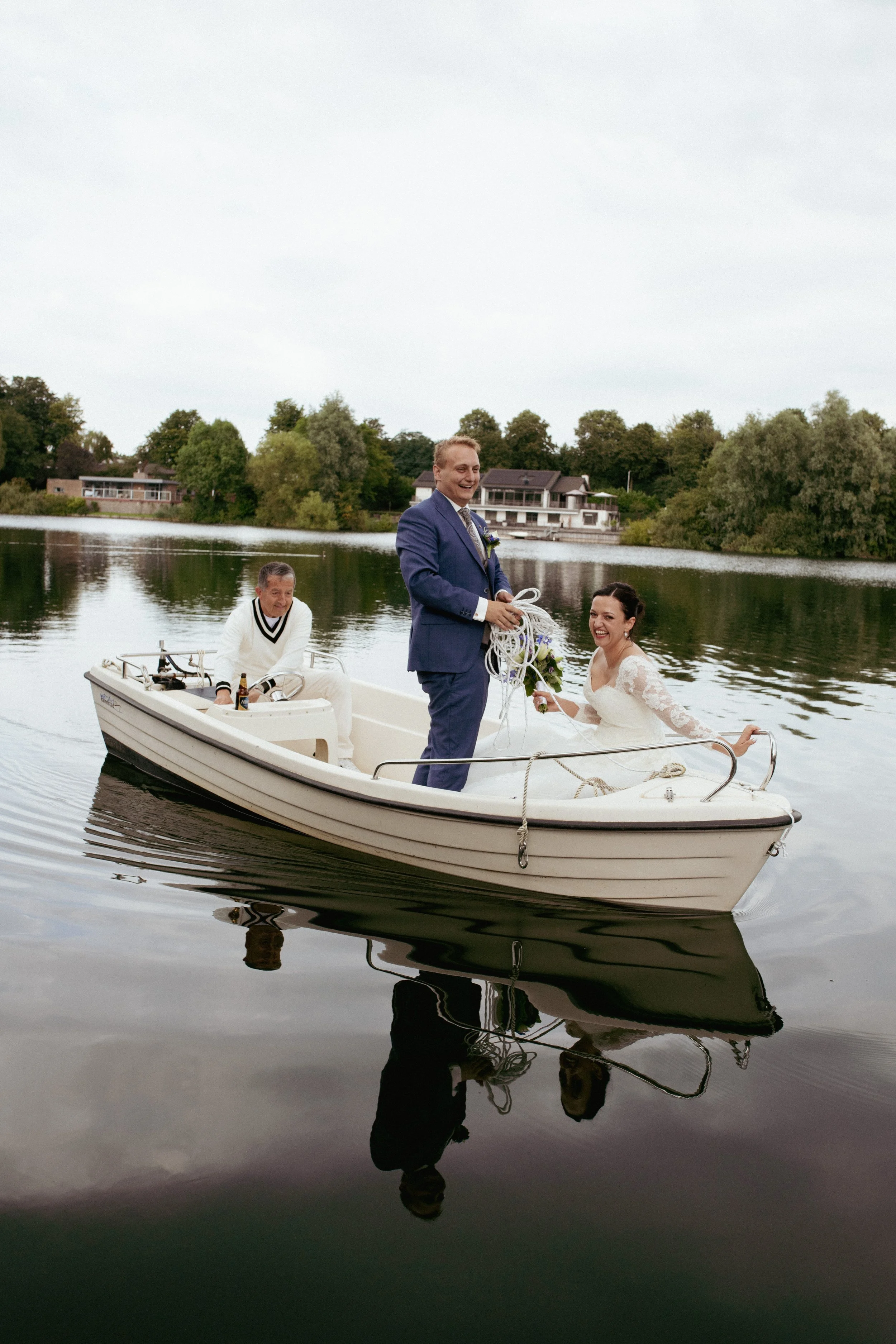 Fotografie op bruiloft. Een bruid en bruidegom op een boot op het water, met een man die de boot bestuurt, omringd door huizen en bomen aan de oever. Door De Kraamfotograaf.