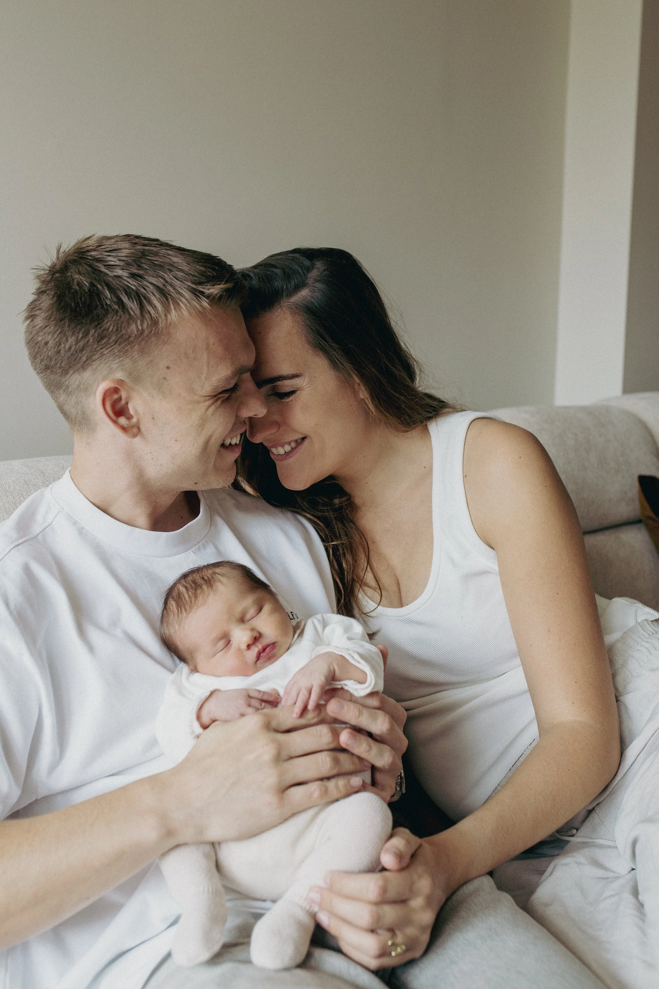 A young couple holding a newborn baby, all smiling and leaning forehead to forehead in a warm, intimate moment.
