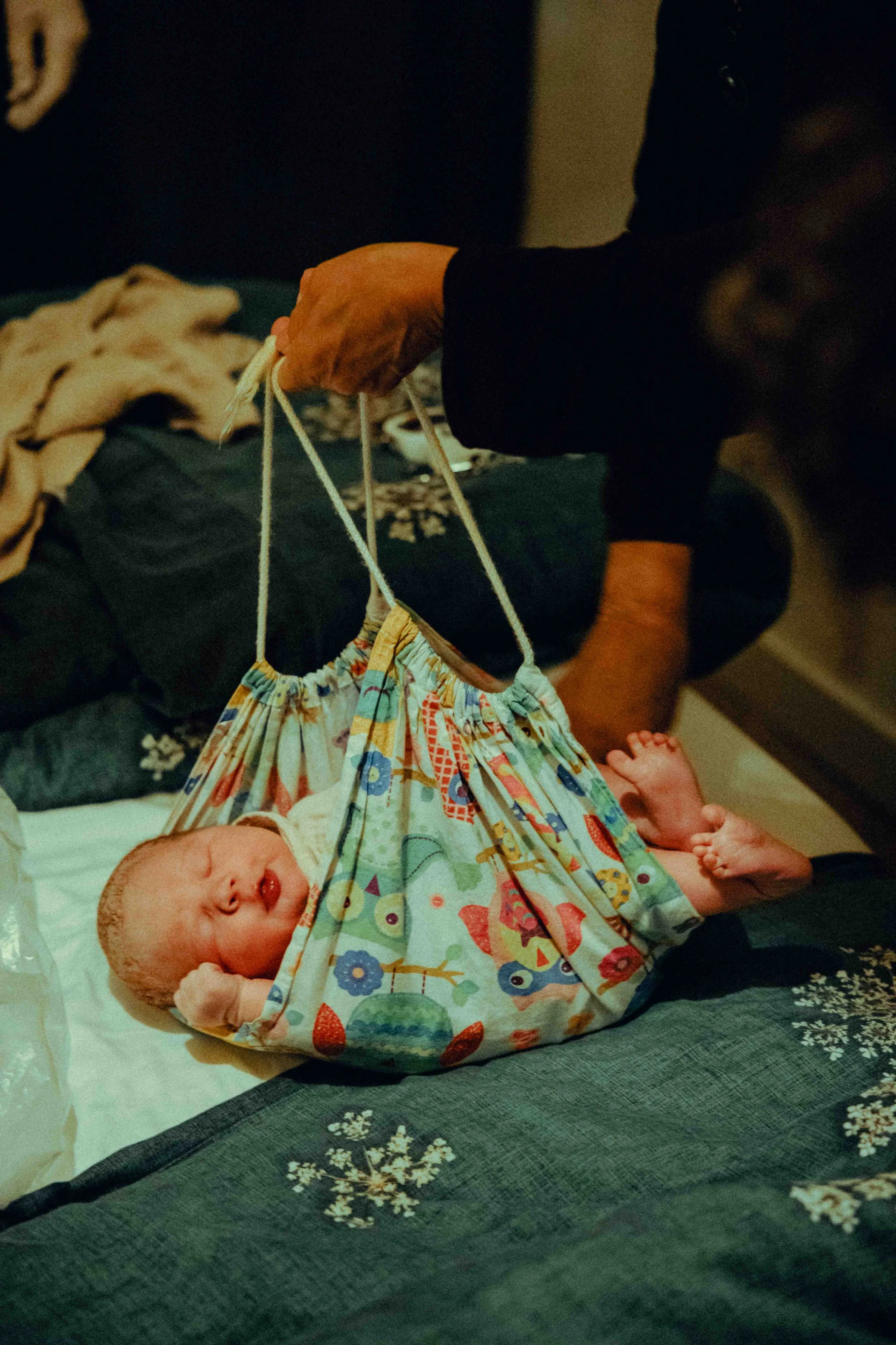 Een pasgeboren baby ligt op een bed, gewikkeld in een kleurrijk luier met dierenpatroon. Een persoon houdt het kind voorzichtig vast.    Geboortefotografie door Maud Fernhout (de Kraamfotograaf).