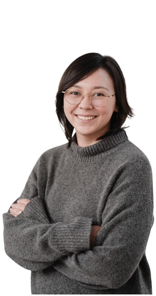A young woman with short dark hair, glasses, and a gray sweater is smiling with arms crossed against a plain white background.