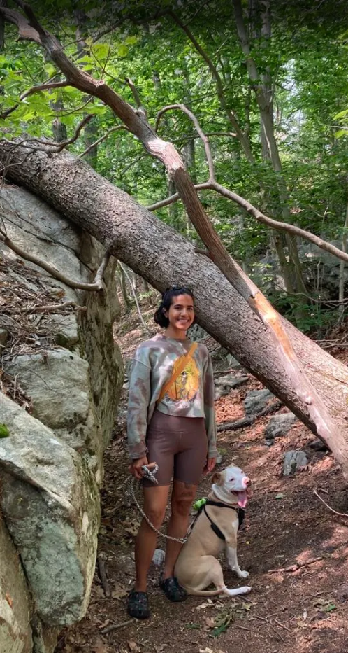 A woman standing on a wooded trail with a large dog sitting beside her, surrounded by green trees and rocks, with a fallen tree leaning overhead.
