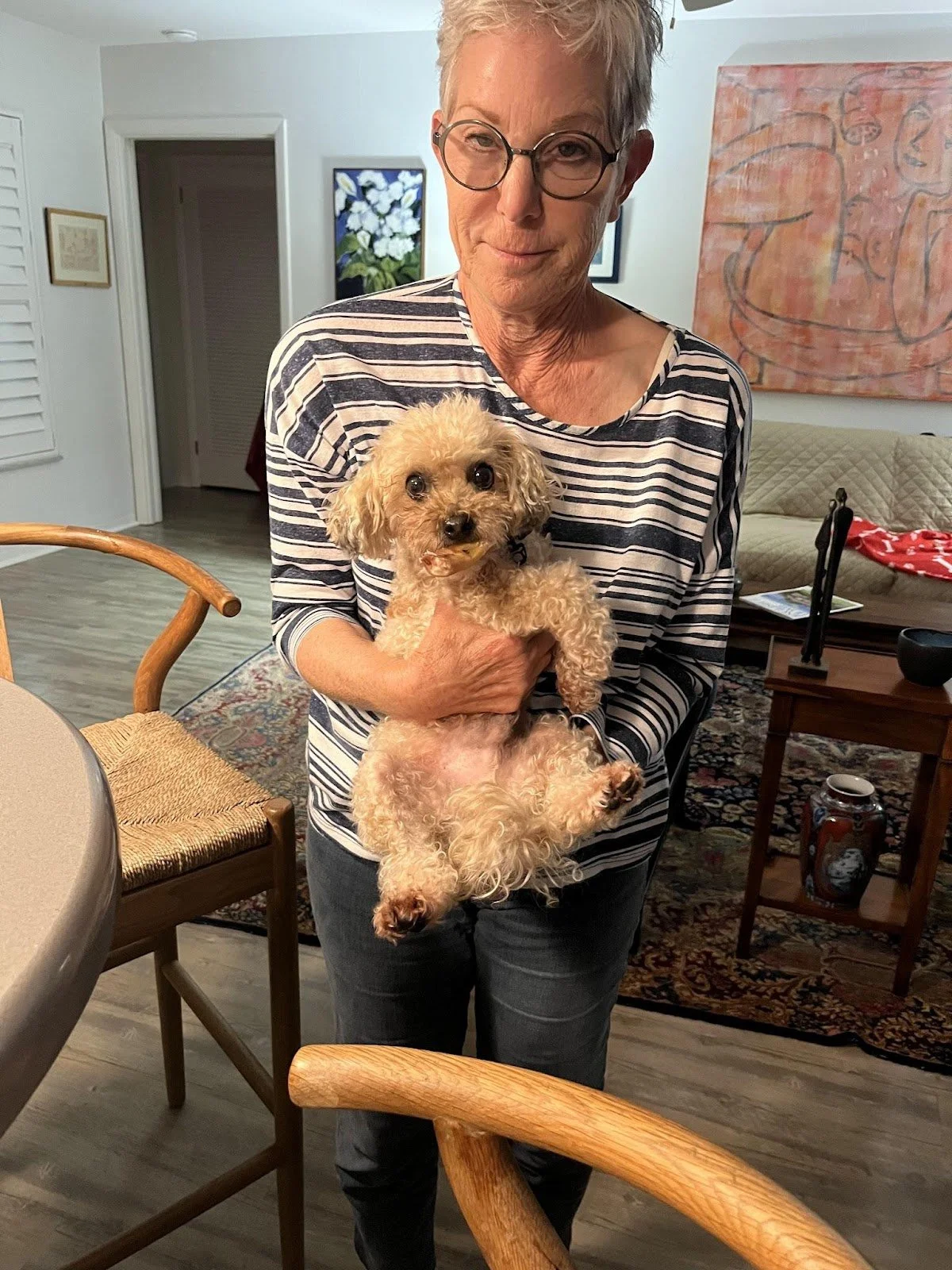An elderly woman with short gray hair and glasses holding a small tan poodle in a cozy living room.