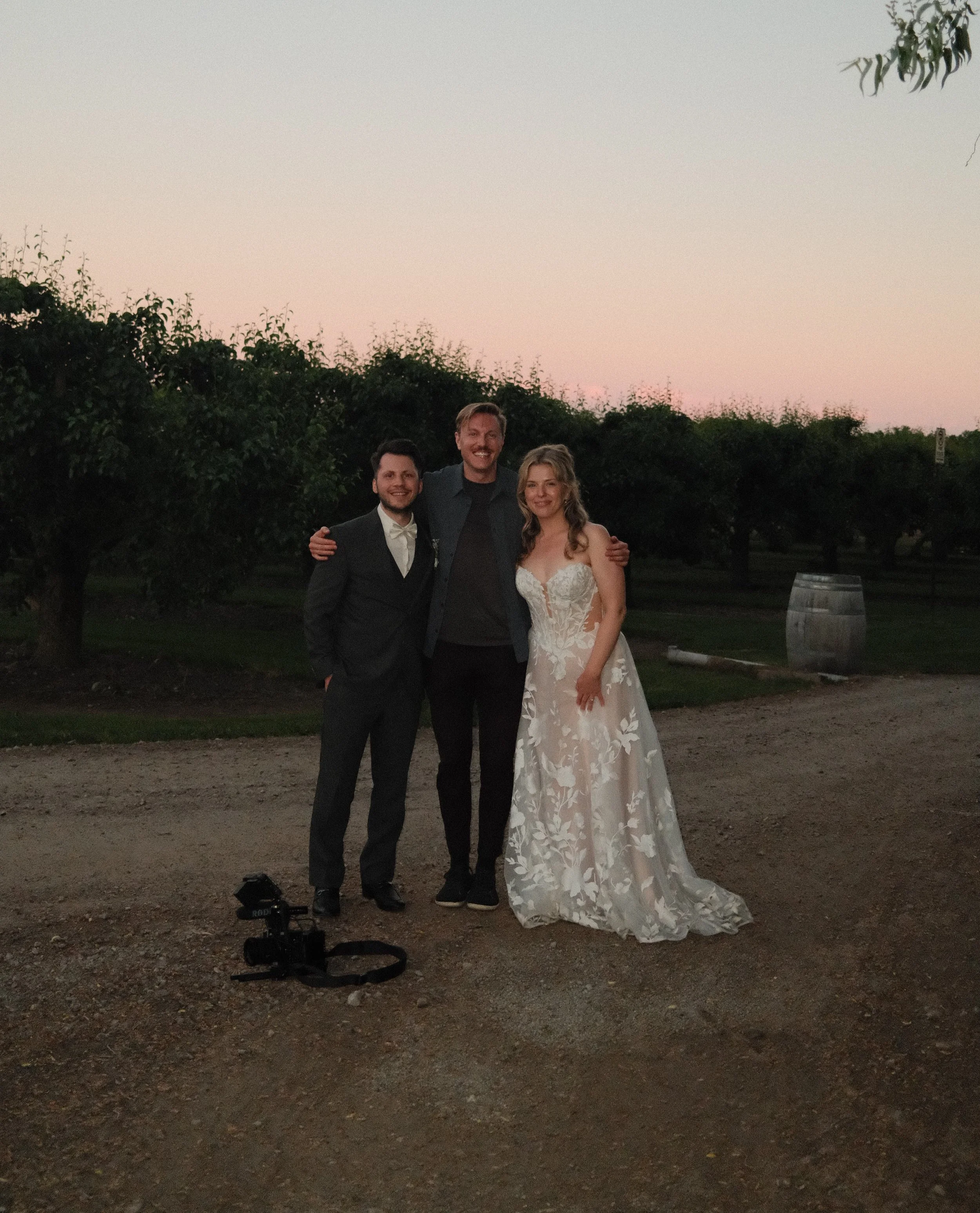 Three people standing outdoors at dusk, a man in a tuxedo, another man in a dress shirt and jeans, and a woman in a white wedding dress. They are smiling and posing closely together, with trees and a barrel in the background and a camera on the ground in front of them.