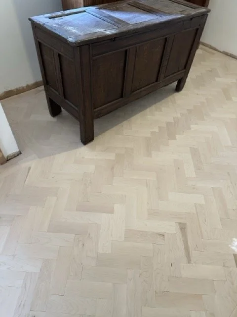 A dark wooden chest with a weathered top sits against a wall in a room with light hardwood herringbone flooring.