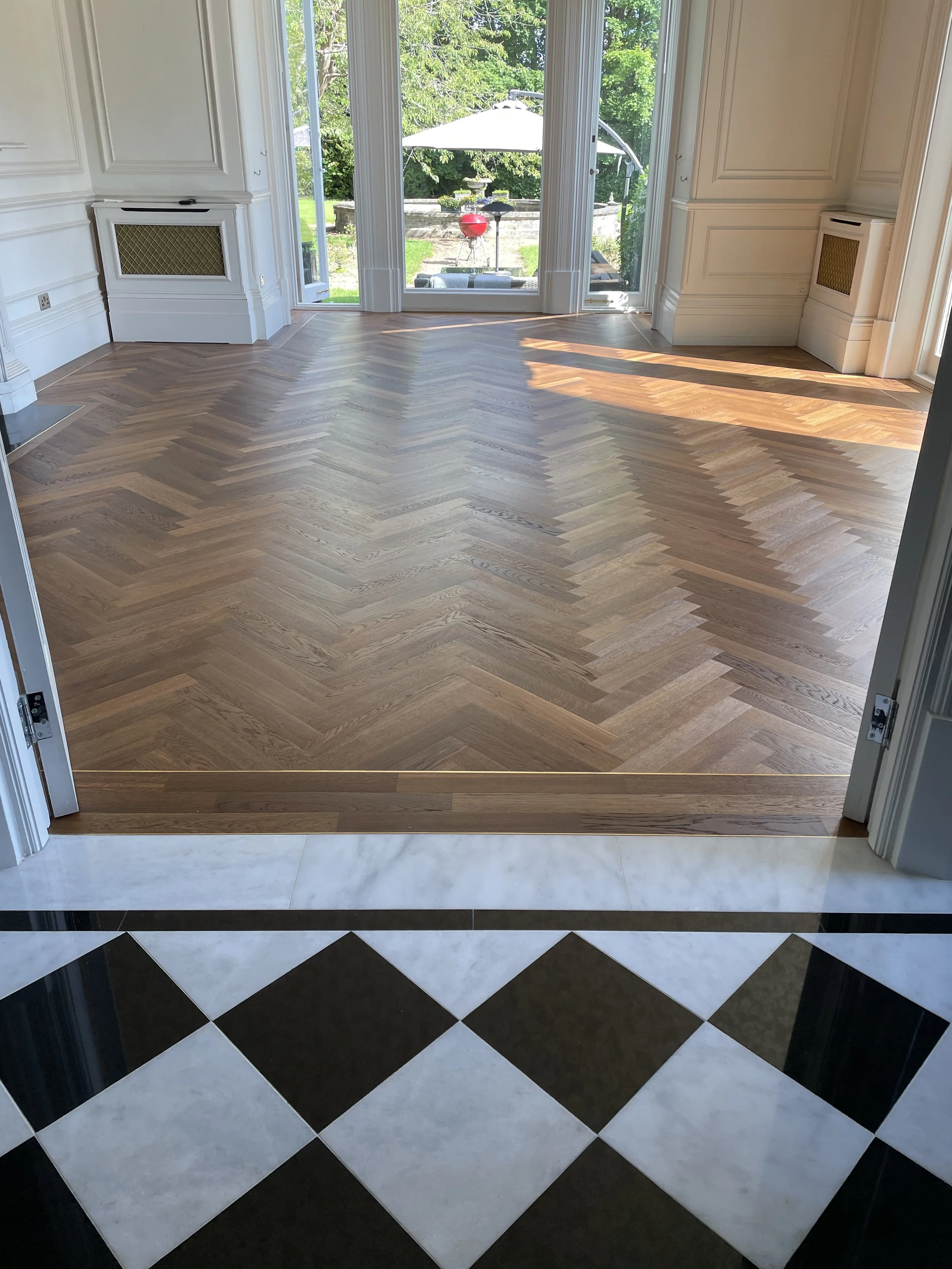View from an interior doorway showing a transition from a black-and-white checkered tile floor to a wooden floor leading to a sunlit room with large windows and a backyard with greenery and outdoor furniture.
