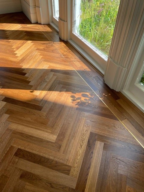 Close-up of a wooden herringbone parquet floor near a glass door, with a measuring tape stretched across the floor indicating its length, sunlight and shadow patterns visible.