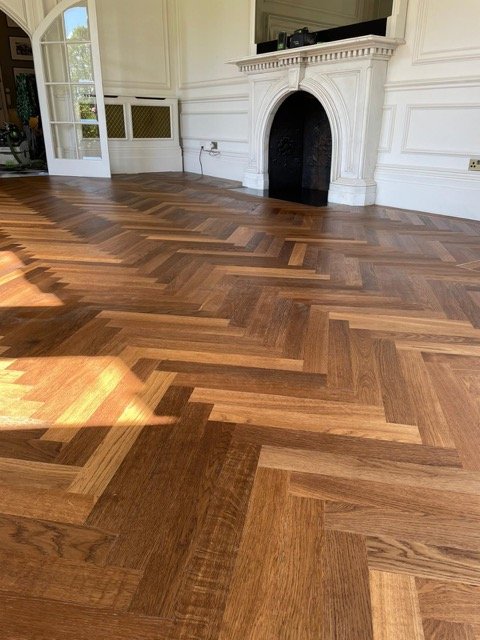 Interior of a room with a herringbone parquet hardwood floor, white paneled walls, a white fireplace with decorative moldings, and a large glass door leading outside.