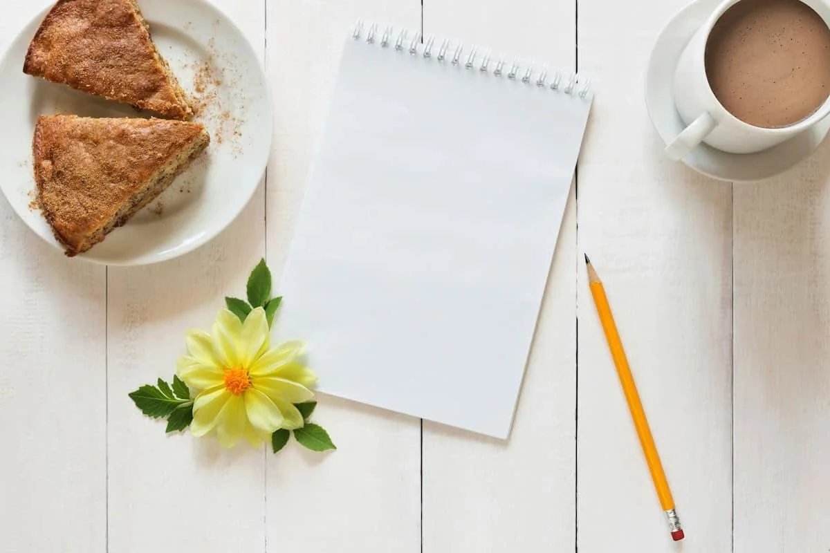 Ein Schreibblock, Bleistift, eine gelbe Blüte und Genuss mit Kuchen und Kaffee