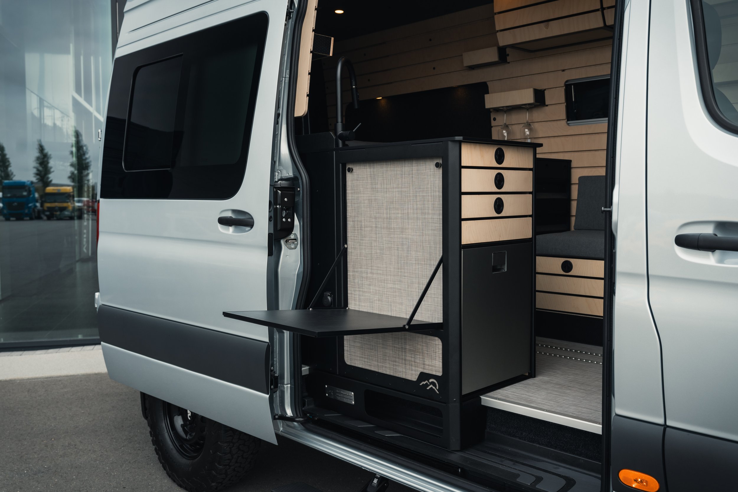 The open side door of a motorhome reveals a fully equipped kitchen inside, including a sink, cabinets, and a bench.