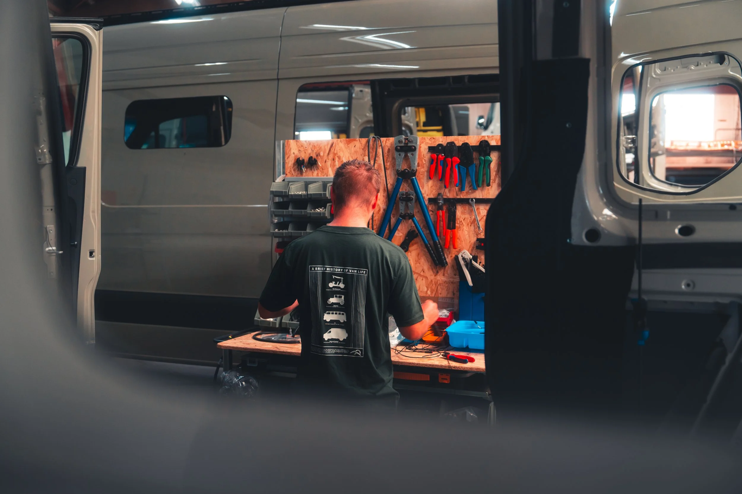 A man is working at a tool rack inside a van, using tools such as screwdrivers and pliers, while looking at the tool rack.