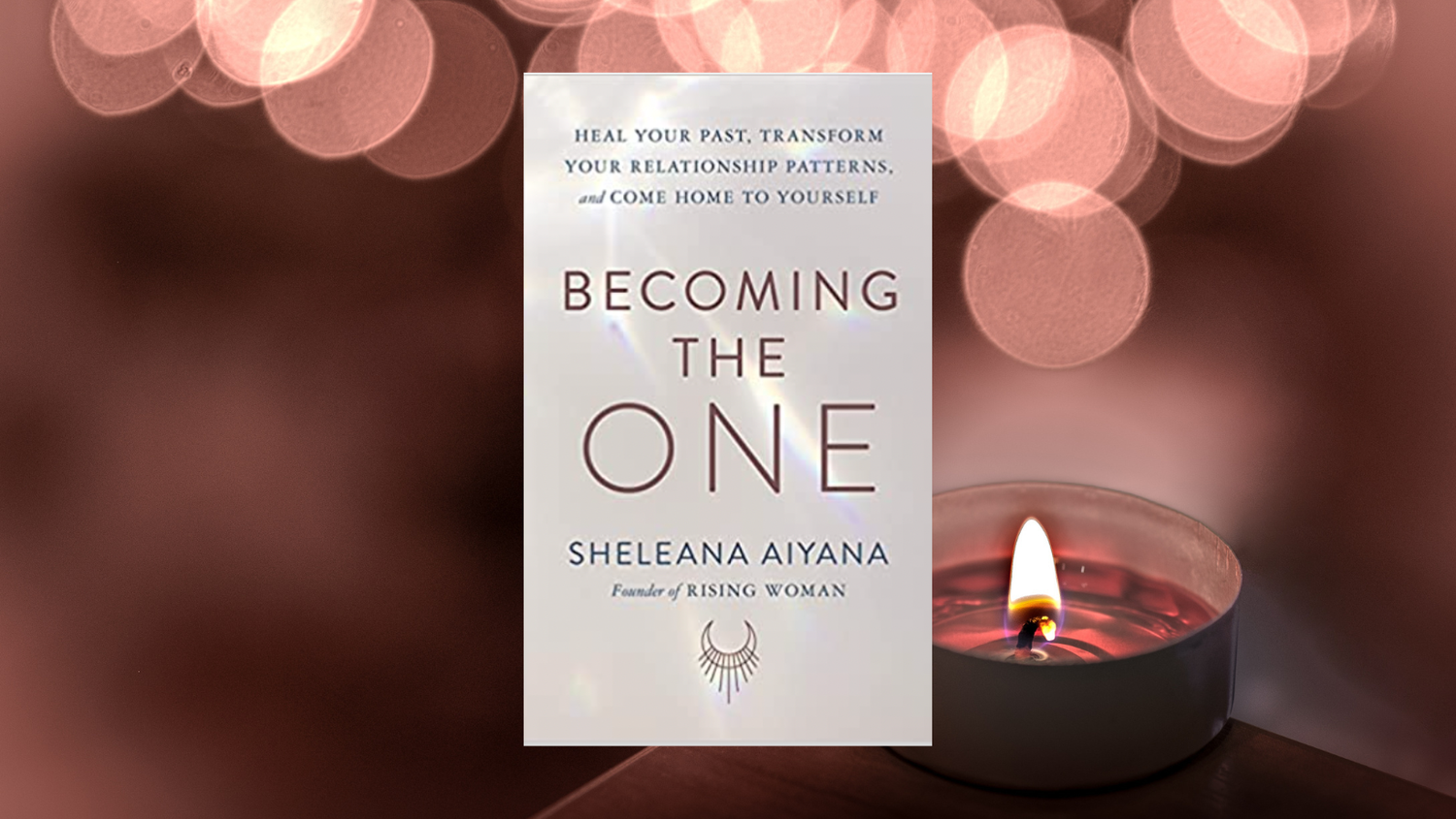 A book titled "Becoming the One" by Sheleena Aiyana stands upright next to a lit candle, with a background of pink and white bokeh lights.