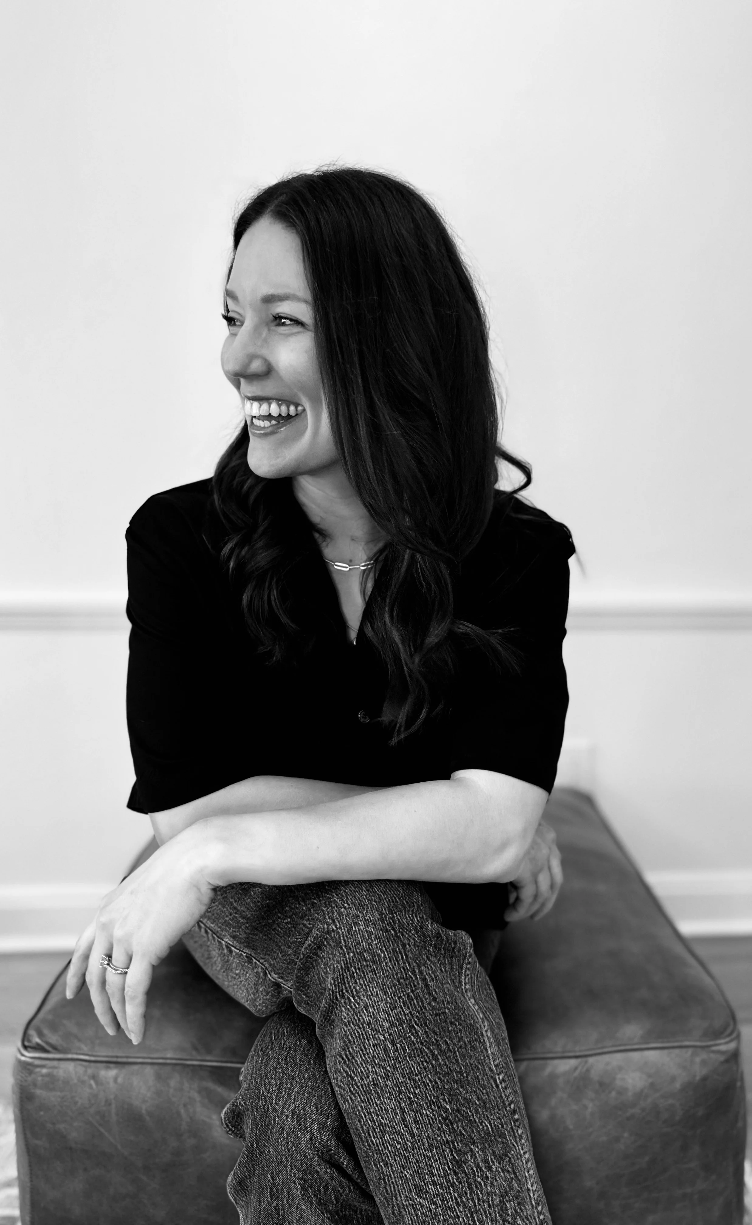 Black and white photo of Tiffaney Marlow sitting on an ottoman, smiling and looking to her left, with her arms crossed and wearing a dark top and jeans.