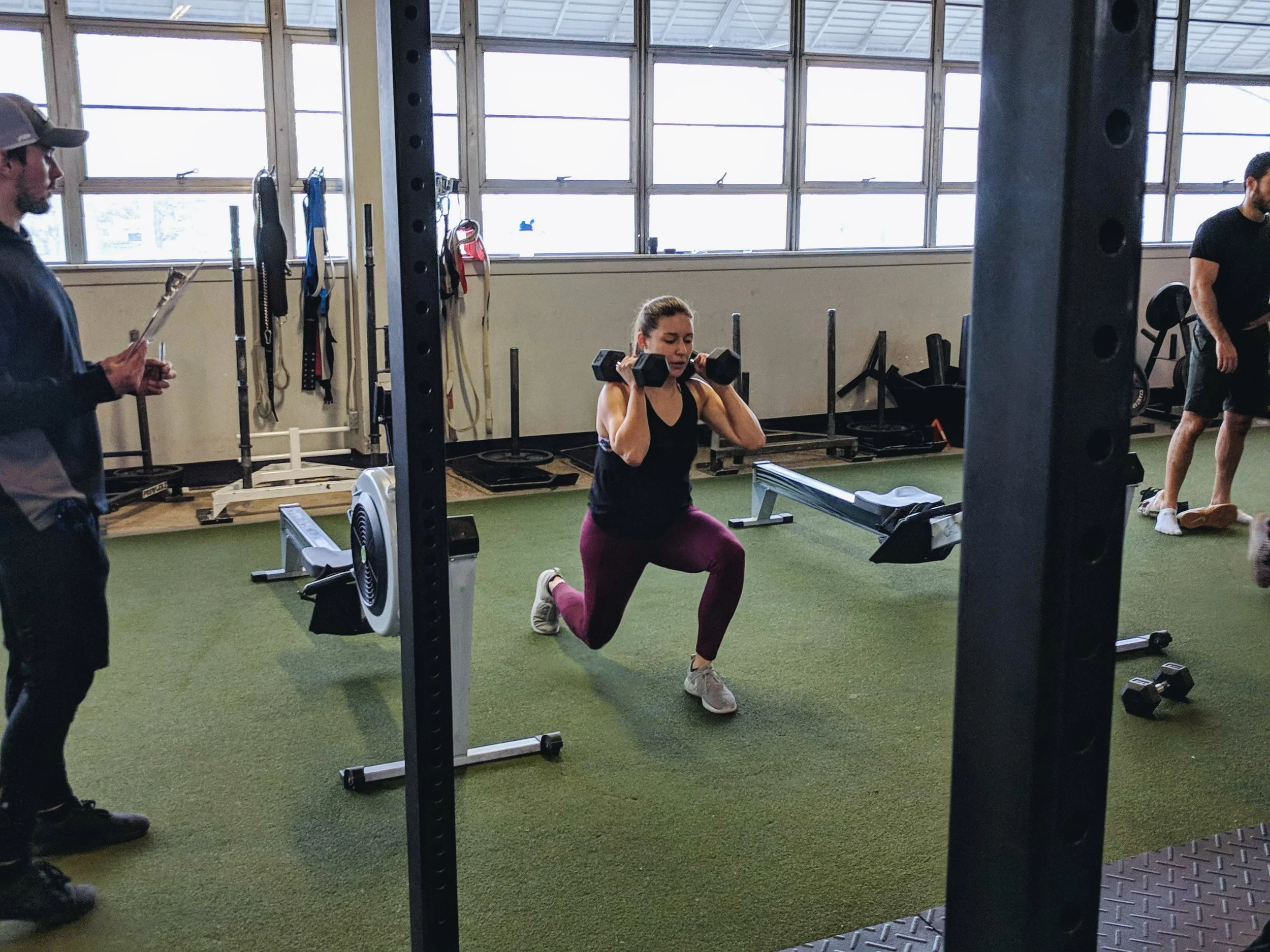 Tiffaney Marlow performing a weighted squat exercise in a gym, with two men nearby.