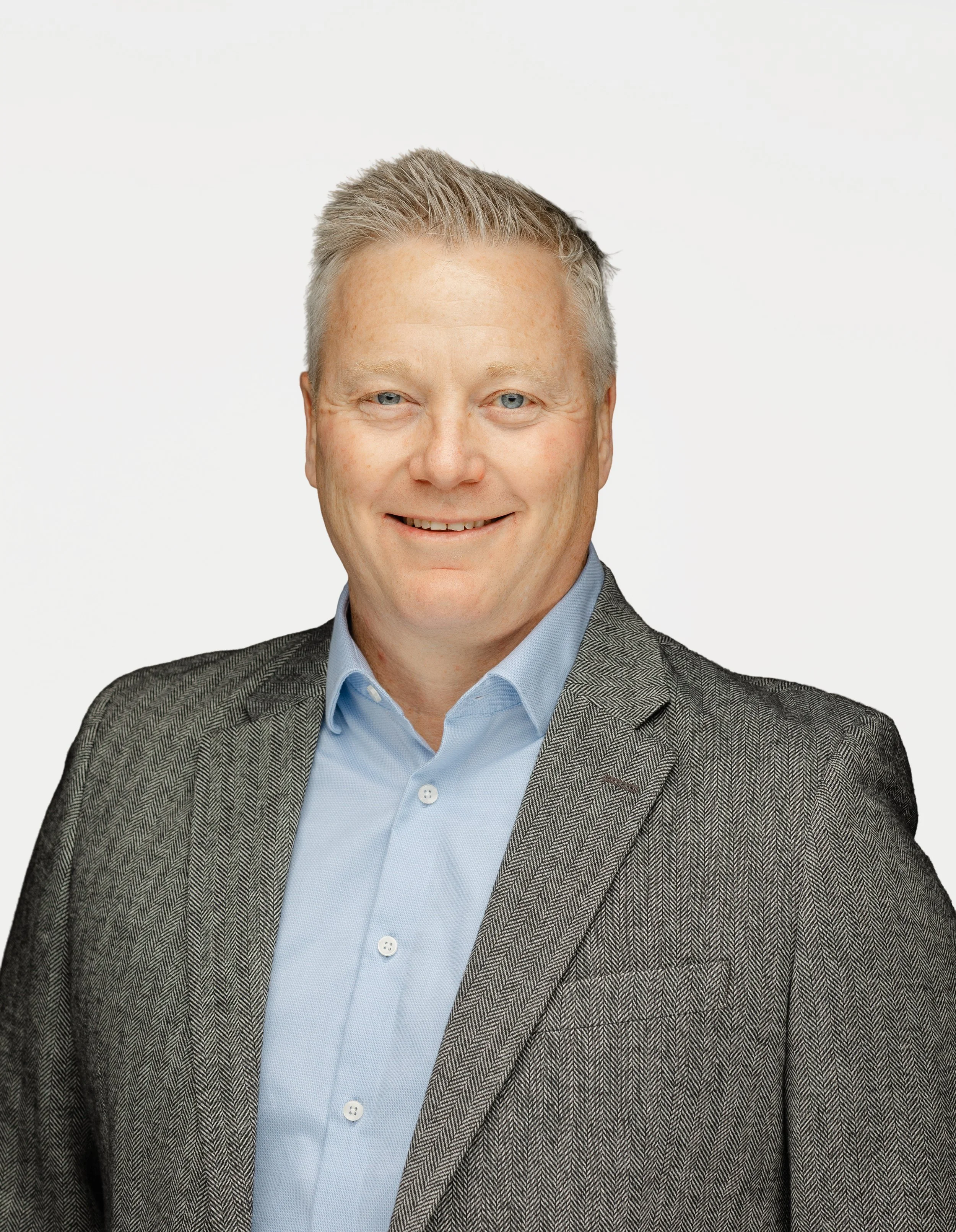 Mission Omega's Head of Advisory, Brad Crall, wearing a gray herringbone blazer and light blue dress shirt, smiling against a white background.