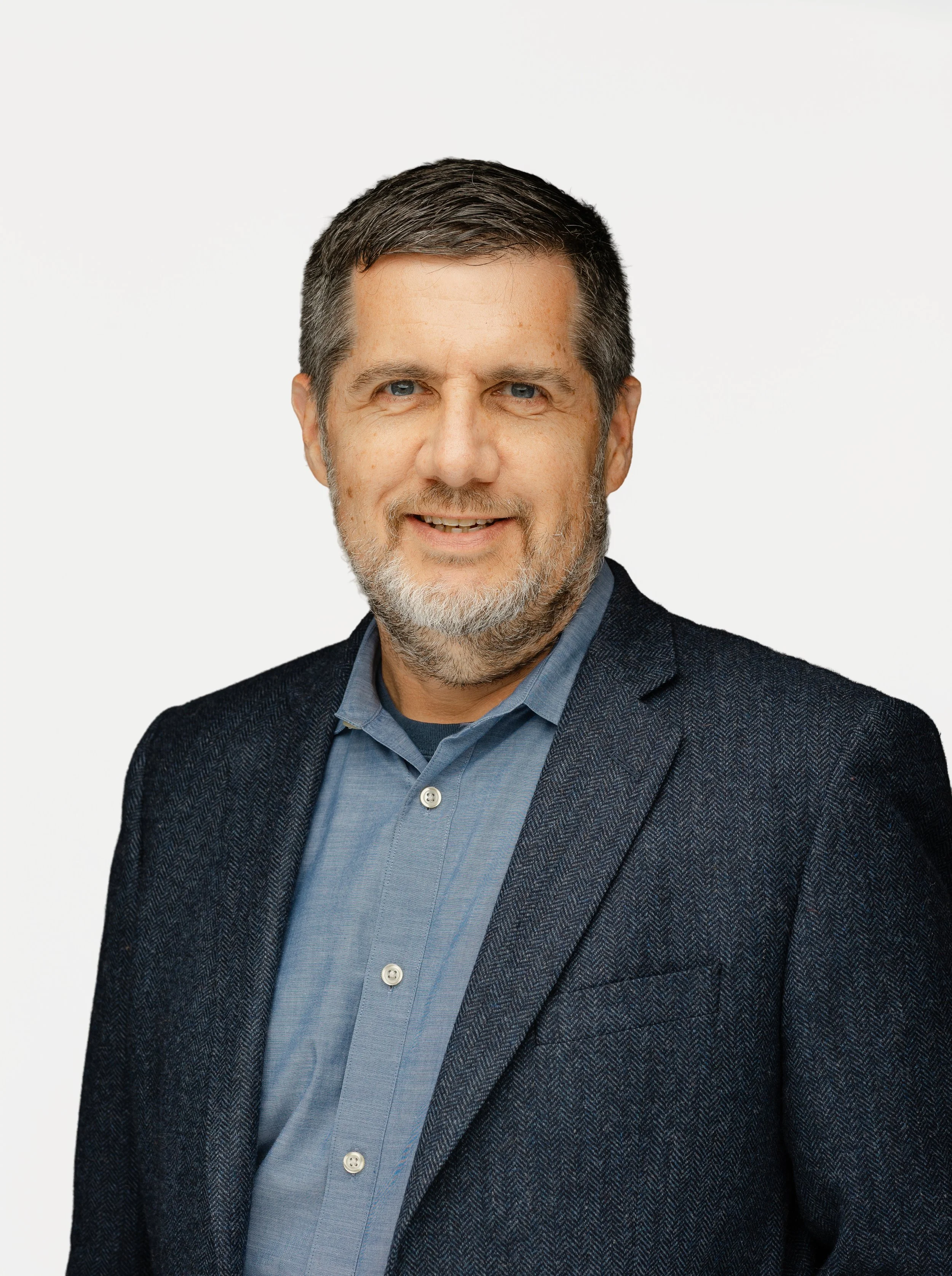 Mission Omega's COO, Tim Chambers, wearing a dark blazer and a blue dress shirt, smiling in front of a plain white background.