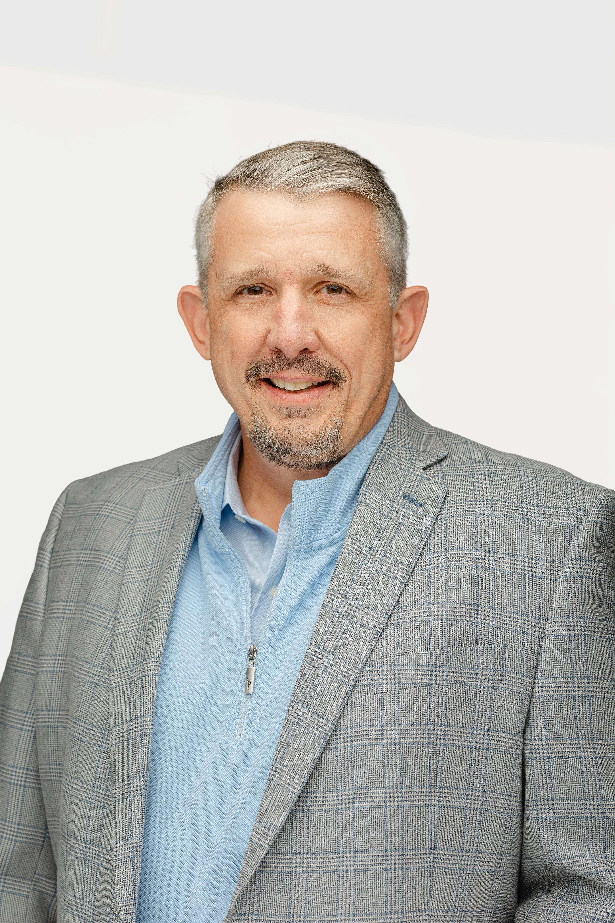 Mission Omega's CEO, Ben Wallach, dressed in a gray checkered blazer and a light blue shirt, smiling at the camera against a plain white background.