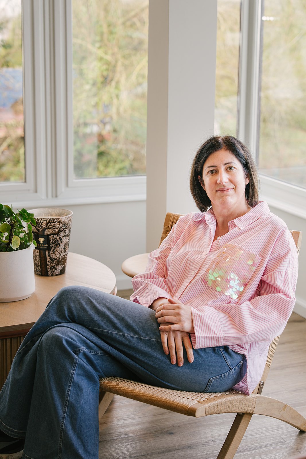 Portret van een vrouw met bruin haar die naar de camera kijkt, zittend tegen een lichte achtergrond, gekleed in een beige trui en blauwe jeans.