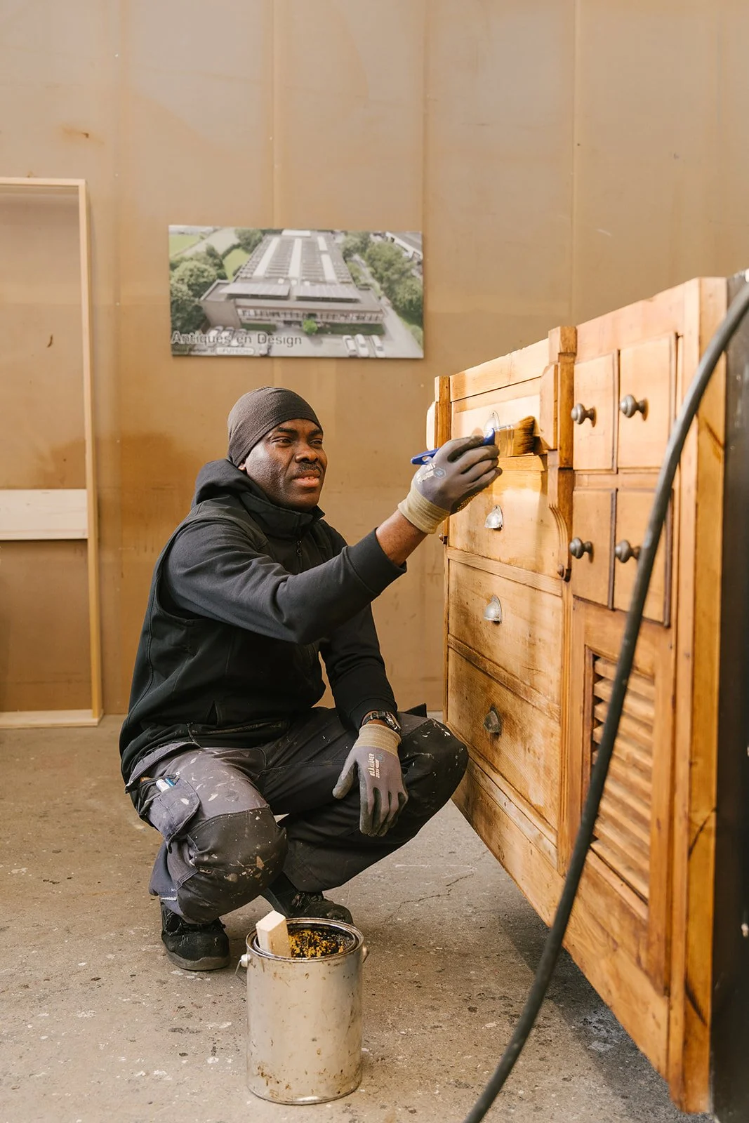 Man met een donkere huid, gekleed in een rood shirt en een donkerbruin muts, met gekruiste armen, staand voor een lichte achtergrond.