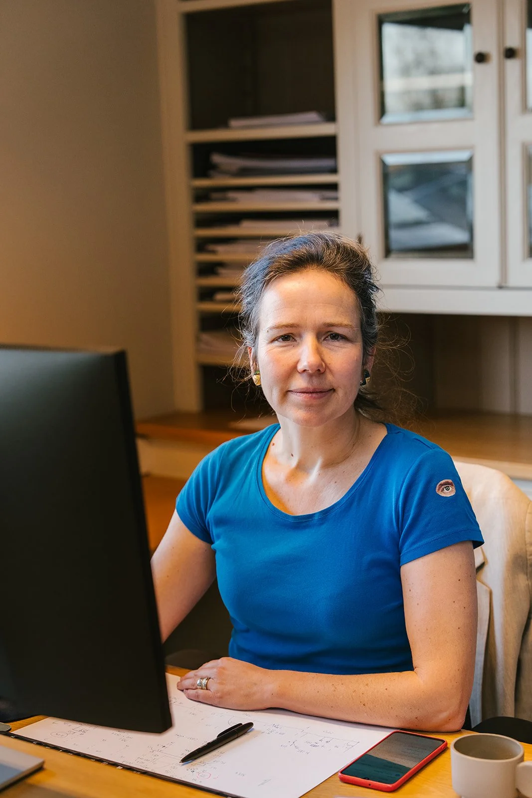 Een vrouw met bruin haar in een lage paardenstaart, lachend, draagt een blauwe gehaakte trui en een witte top, tegen een lichte achtergrond.