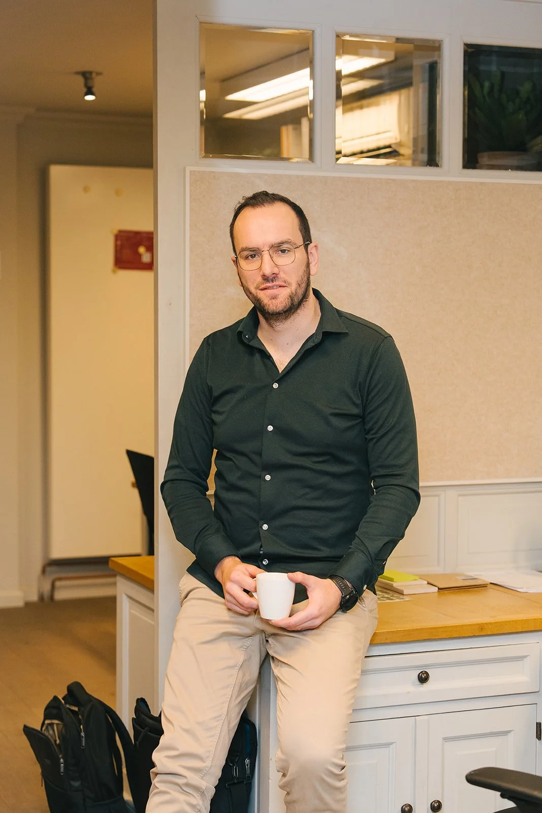 Portretfoto van een jonge man met bruin haar, brilmontuur en een groen overhemd, staand voor een lichte achtergrond.