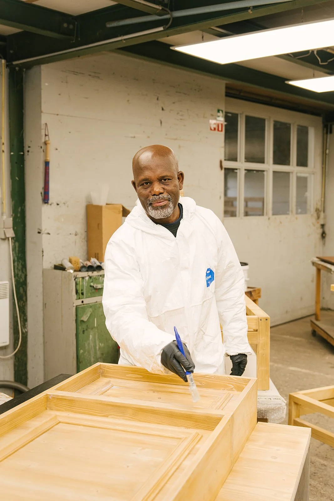 Een man met een donkere huid, grijs baard en bril, gekleed in een donkerroze hoodie, staat tegen een lichte muur en kijkt naar de camera.