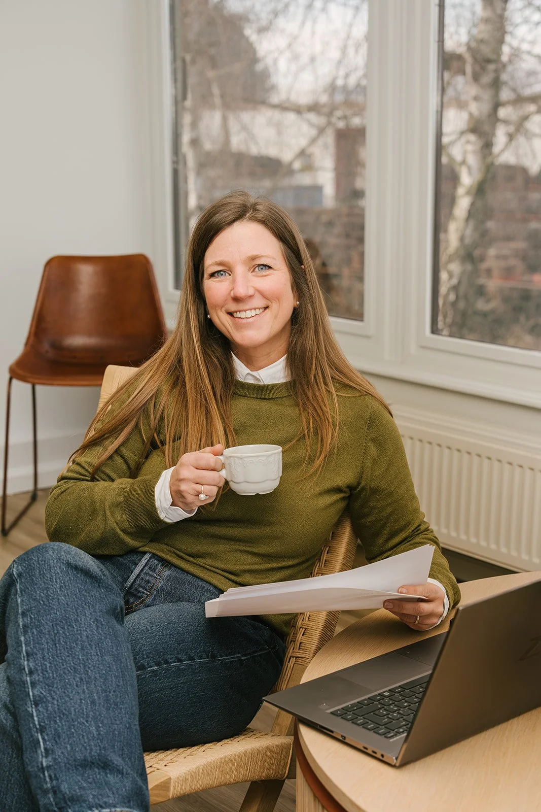 Portret van een lachende vrouw met bruin haar in een zwarte blouse en een donkergekleurd vest, staand voor een lichte, effen achtergrond.