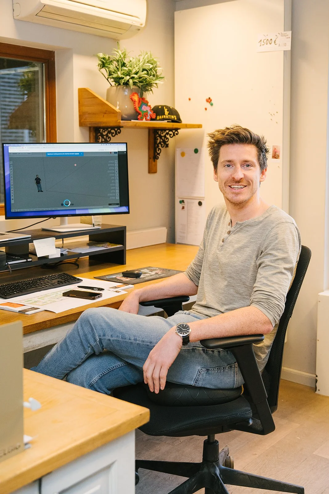 Een man met bruin haar en een glimlach, gekleed in een zwarte trui en een witte blouse, staat voor een lichte muur.