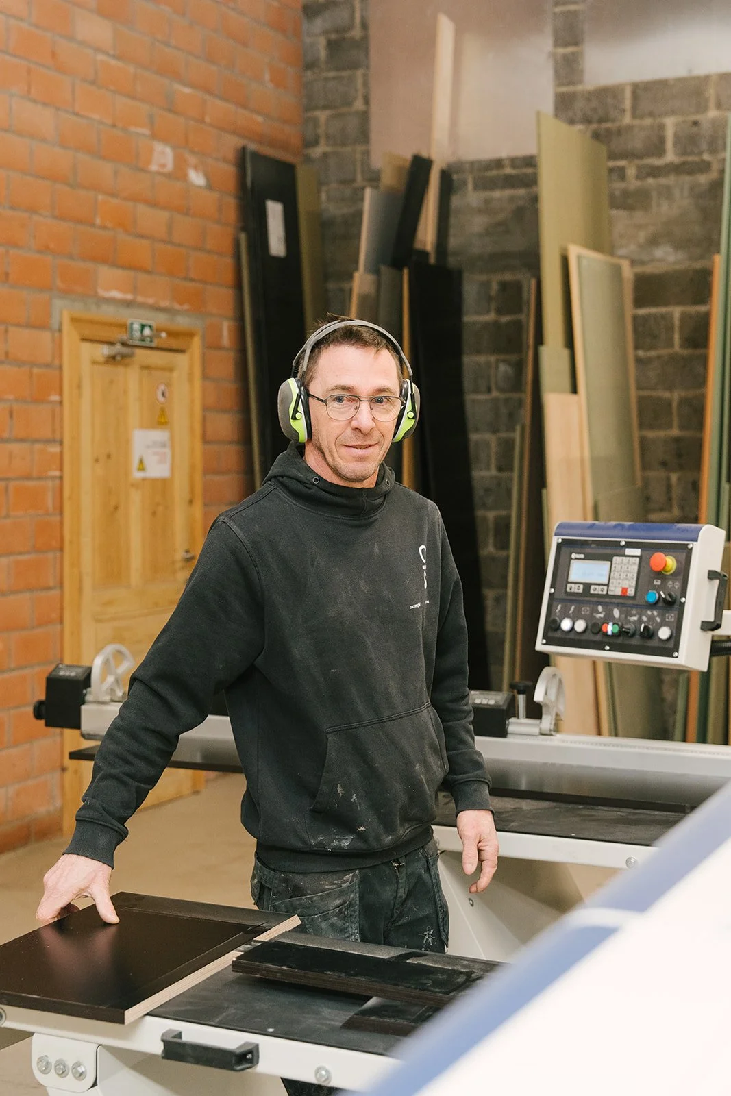 Man met bril en kort haar draagt een donkergroene sweatshirt met de tekst 'Antiques & Design' en staat voor een witte muur.
